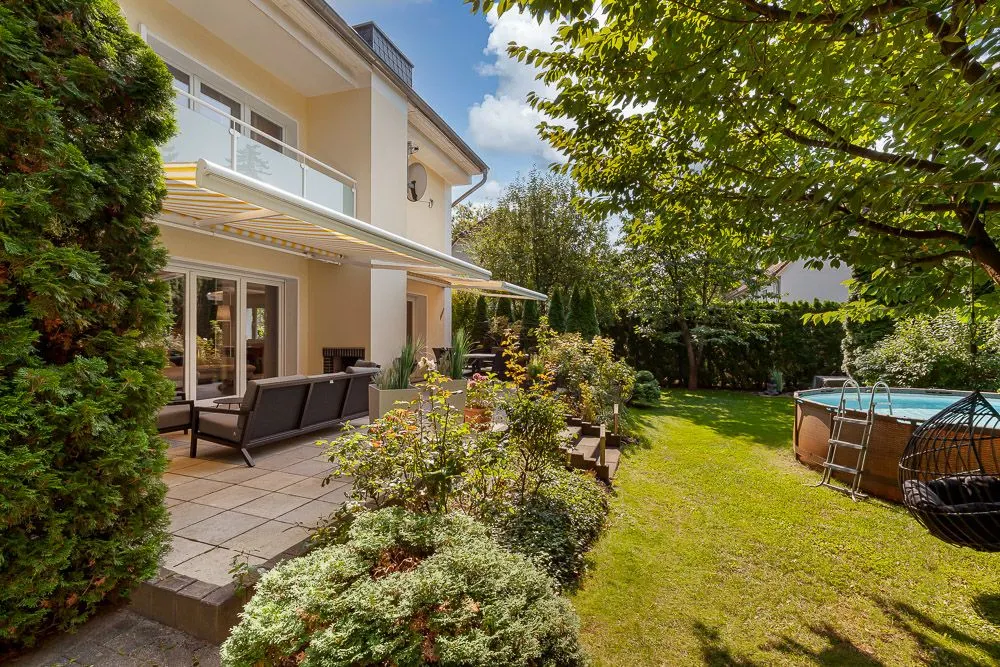 Garten mit Terrasse, Sitzmöbeln, Pflanzen und rundem Pool an einem Wohnhaus bei Sonnenschein.