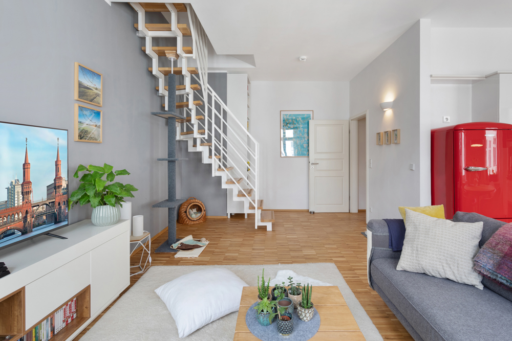 Modernes Wohnzimmer mit grauem Sofa, rotem Kühlschrank und offener Treppe.