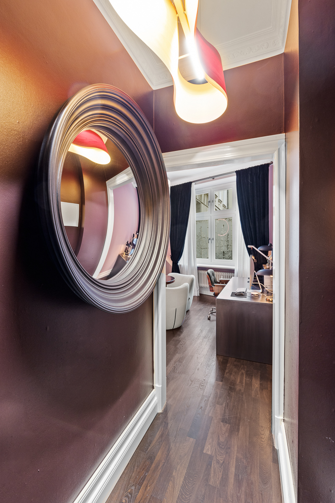 Schmaler Flur mit rundem Spiegel und Blick in ein Zimmer mit Schreibtisch und Sessel.