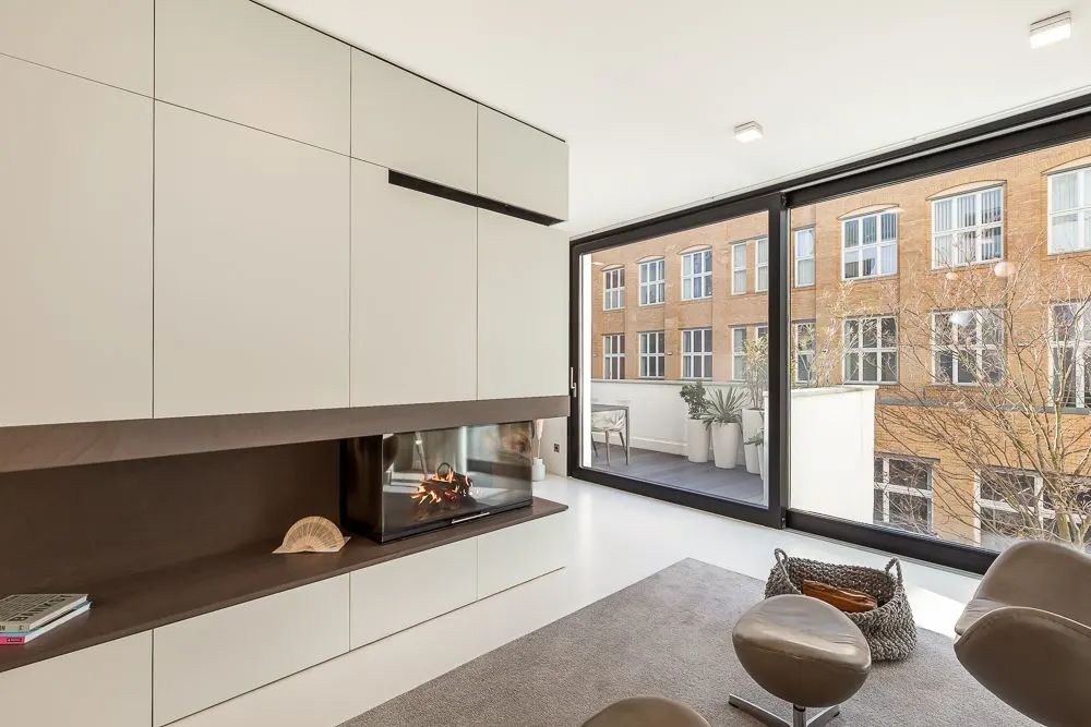 Modernes Wohnzimmer mit Kamin, großen Fenstern und Zugang zu einem Balkon mit Pflanzen.