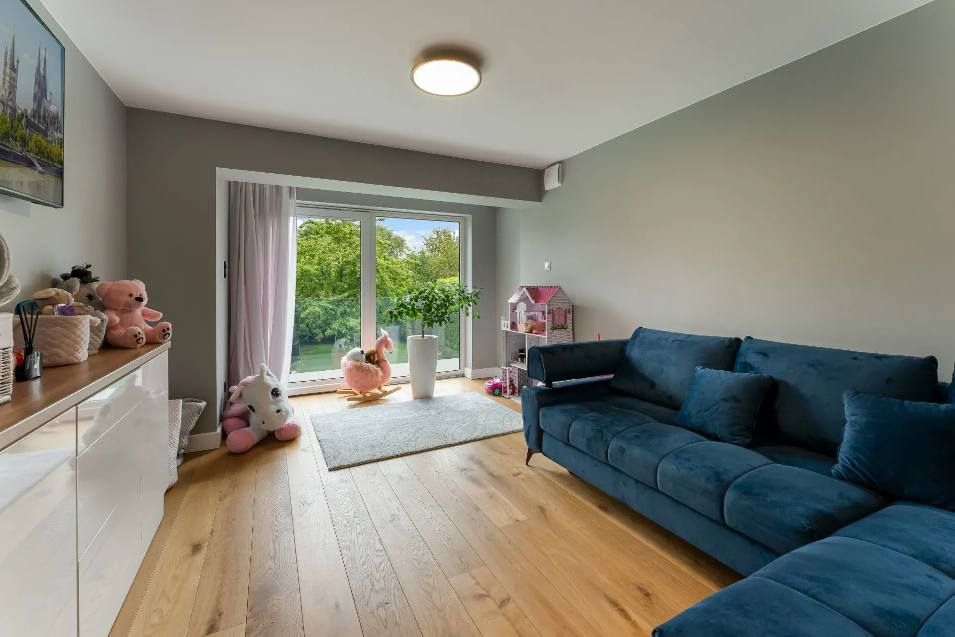 Helles Wohnzimmer mit blauem Sofa, Spielzeug, Holzboden und großem Fenster mit Blick ins Grüne.