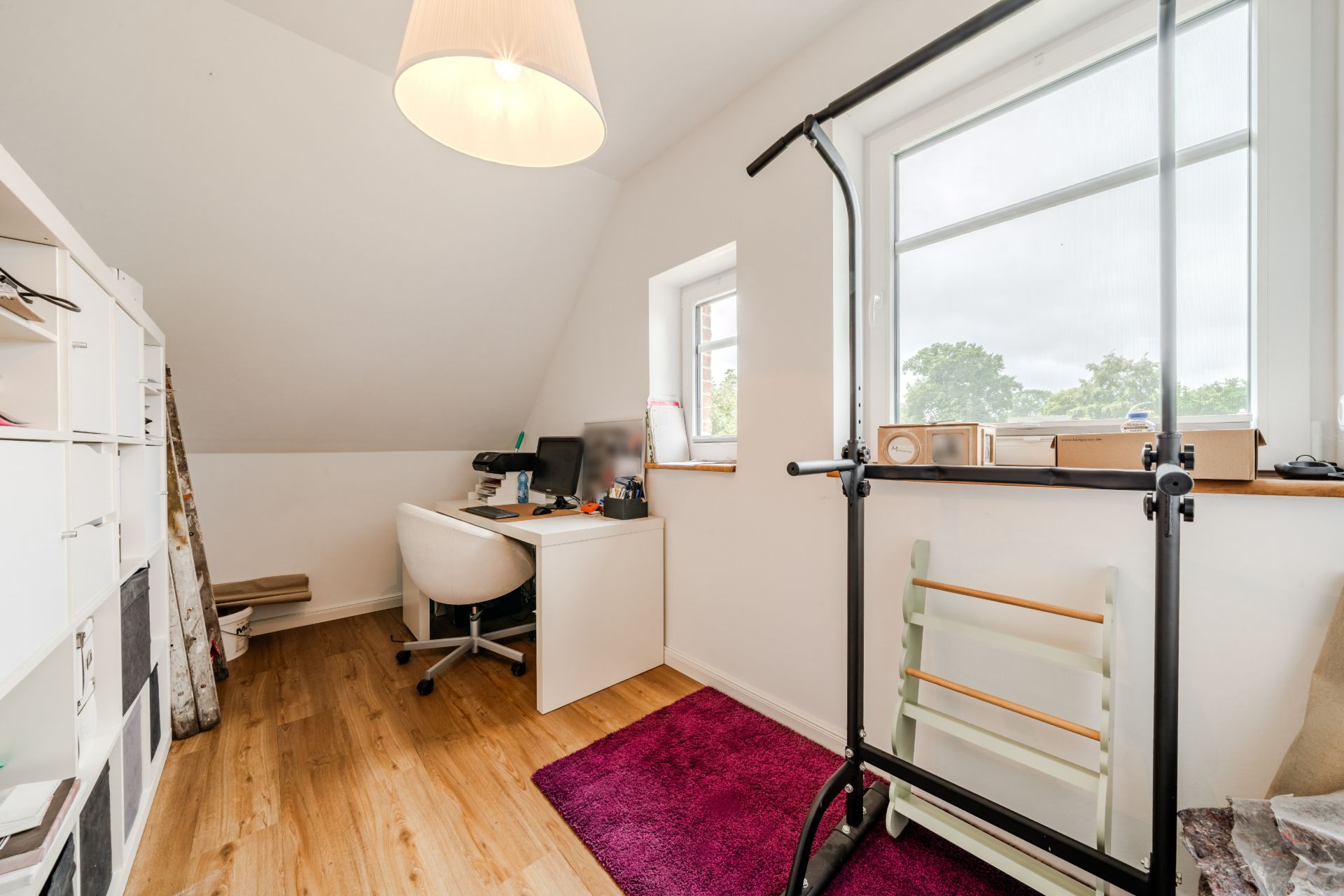 Helles Arbeitszimmer mit Schreibtisch, Bürostuhl, Regal, Fitnessgerät und Teppich auf Holzfußboden.