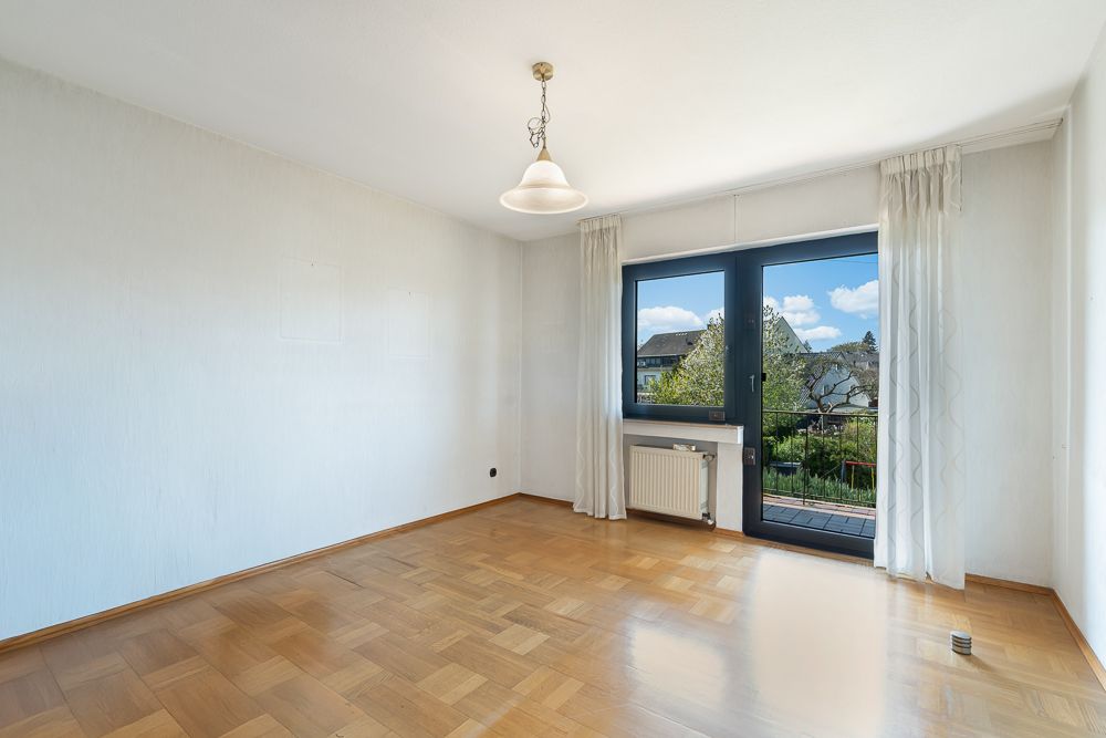 Helles, leeres Zimmer mit Parkettboden, großem Fenster und Glastür zum Balkon.