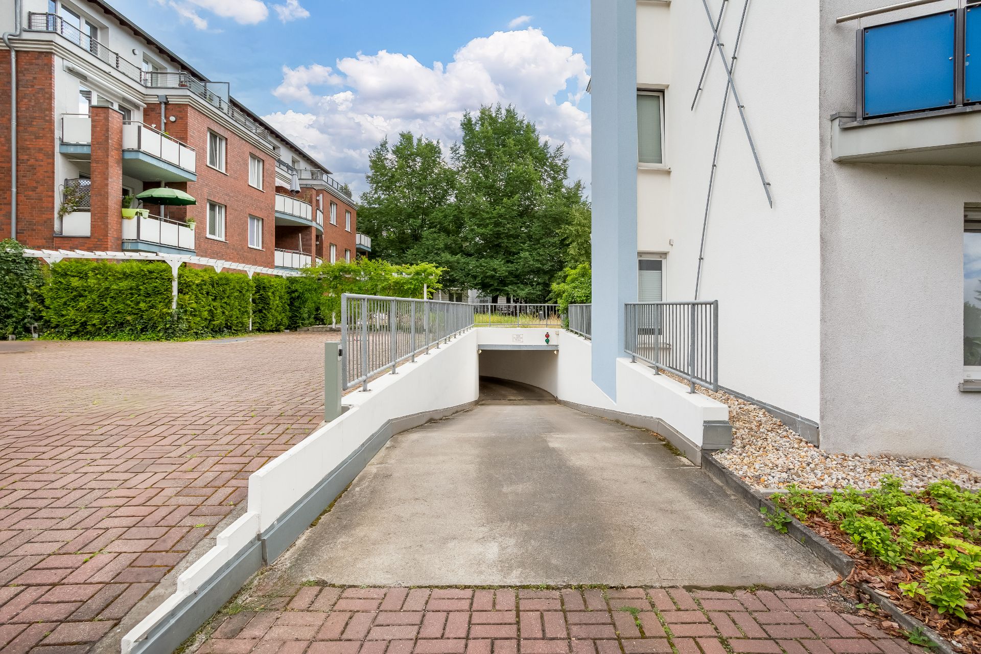 Auffahrt zur Tiefgarage neben einem Wohngebäude mit Bäumen im Hintergrund.