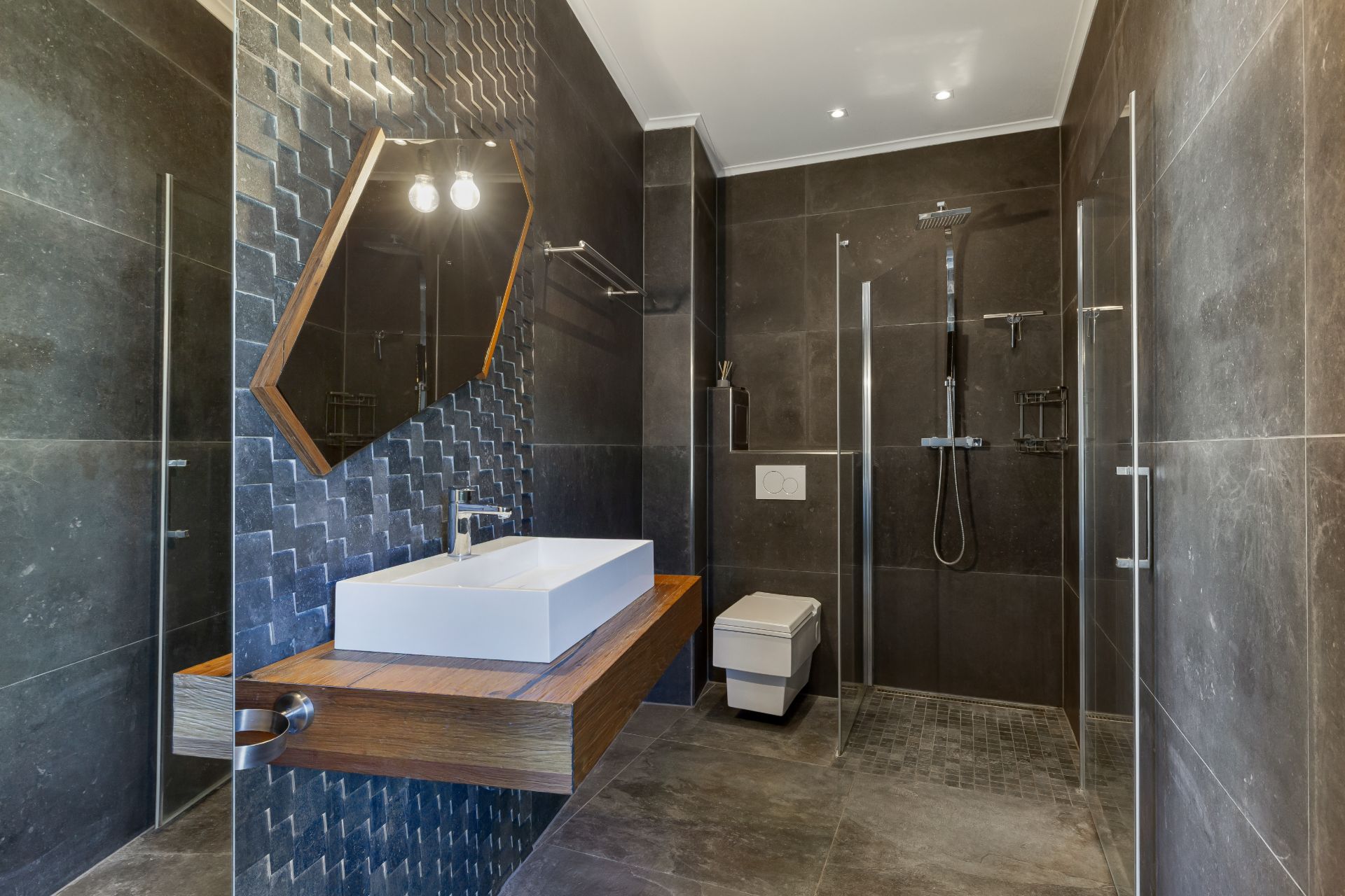 Modernes Bad mit begehbarer Dusche, Waschbecken, Wandspiegel und dunklen Fliesen.
