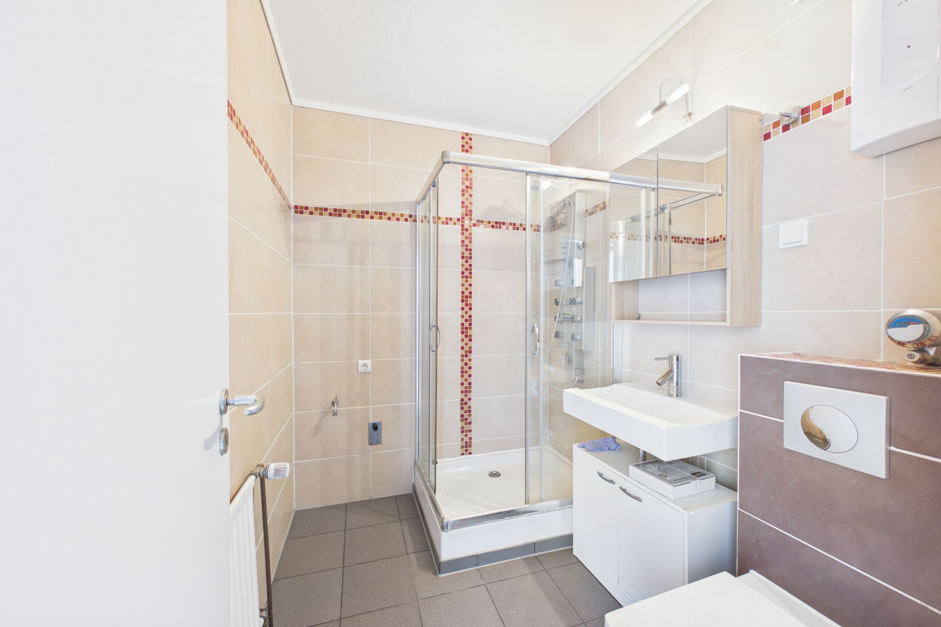Modernes Badezimmer mit Dusche, Waschbecken, Spiegel und WC, beige Fliesen mit roten Akzenten.