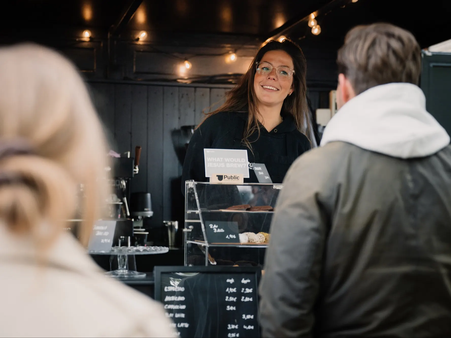 Frau bedient Kunden an einem Kaffeestand mit Gebäck und Getränkekarte.