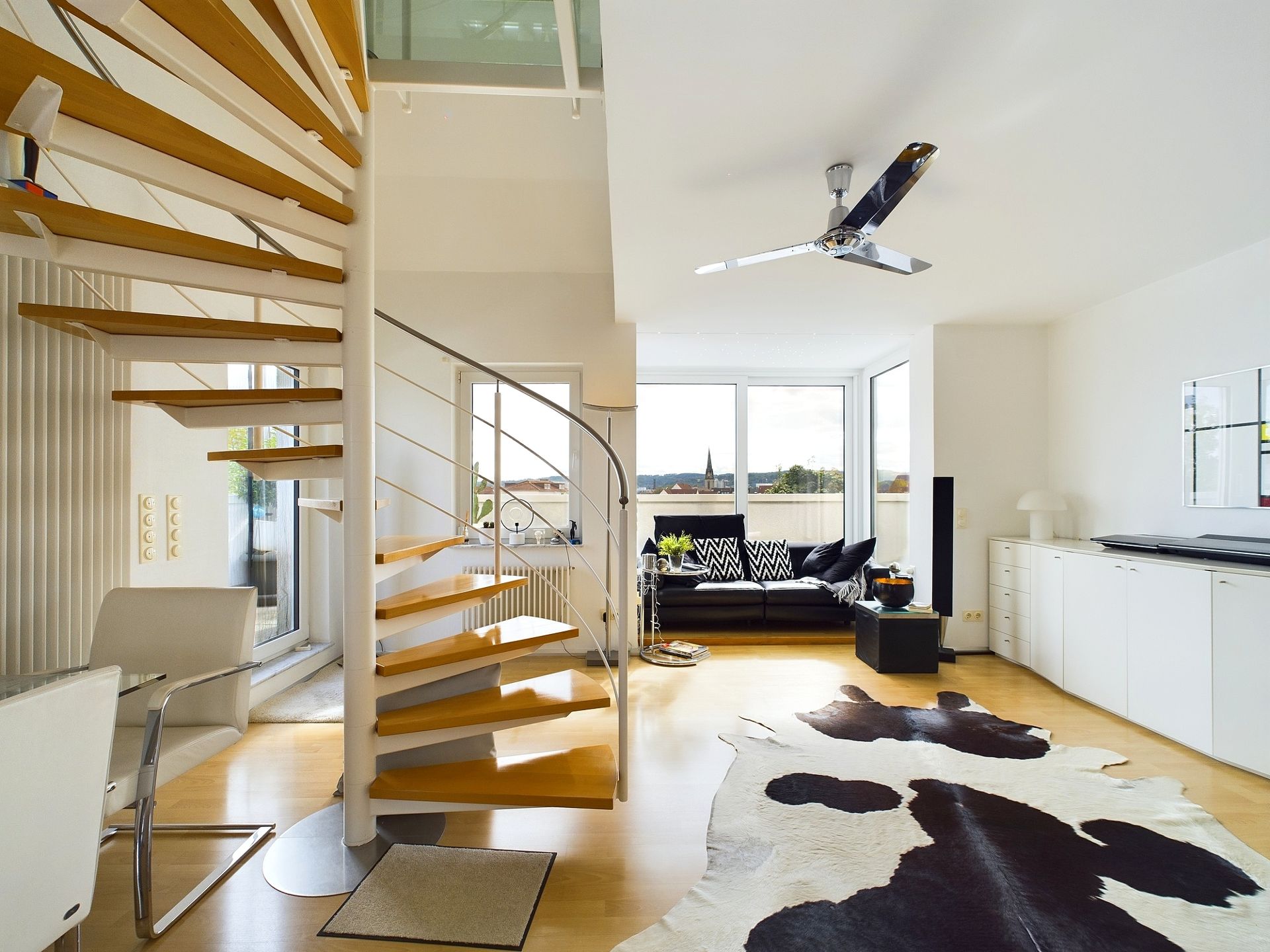 Modernes Wohnzimmer mit Holz-Wendeltreppe, Sofa, Deckenventilator und Kuhfell-Teppich.