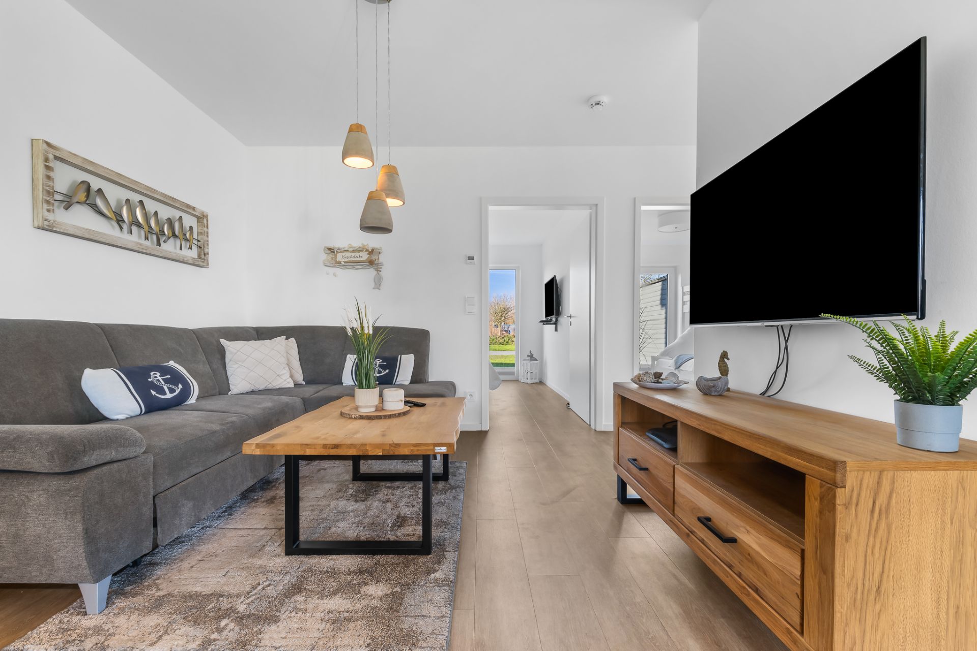 Modernes Wohnzimmer mit grauem Sofa, Holztisch, Fernseher und Pflanzen auf Holzmöbeln.