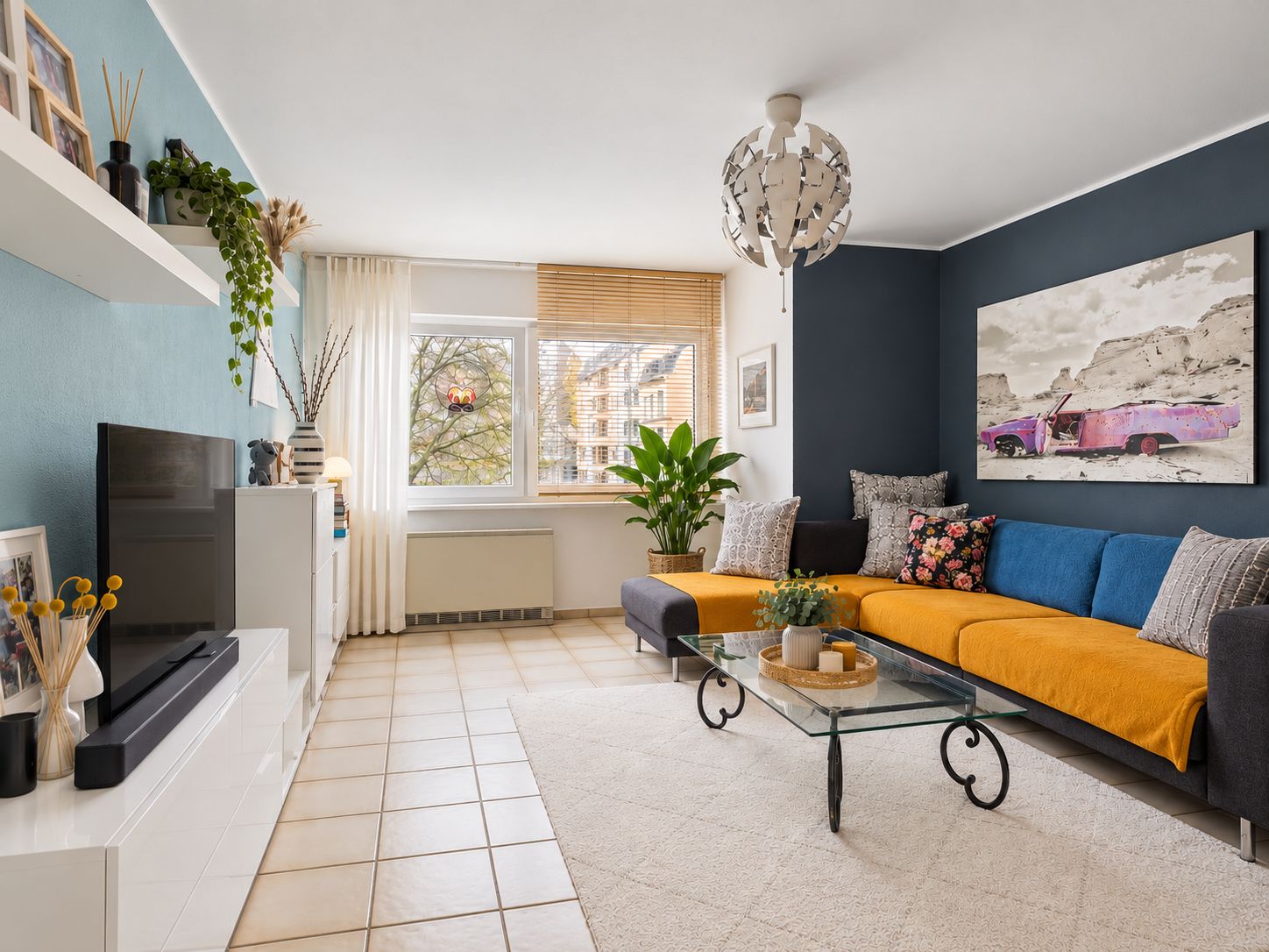 Modernes Wohnzimmer mit gelber Couchdecke, Glastisch und großem Fenster, Pflanzen und Wandbild.