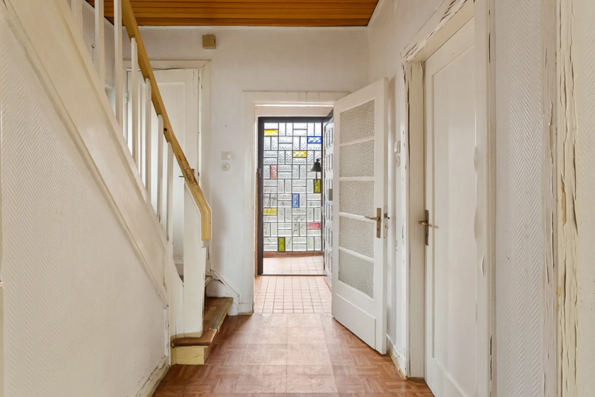 Flur mit Treppe und weißen Türen, am Ende eine Tür mit buntem Glasfenster.