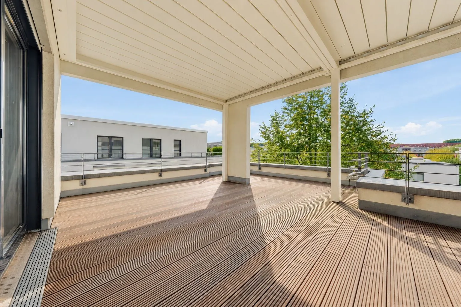 Große überdachte Terrasse mit Holzfußboden und Blick auf Bäume und Nachbargebäude.