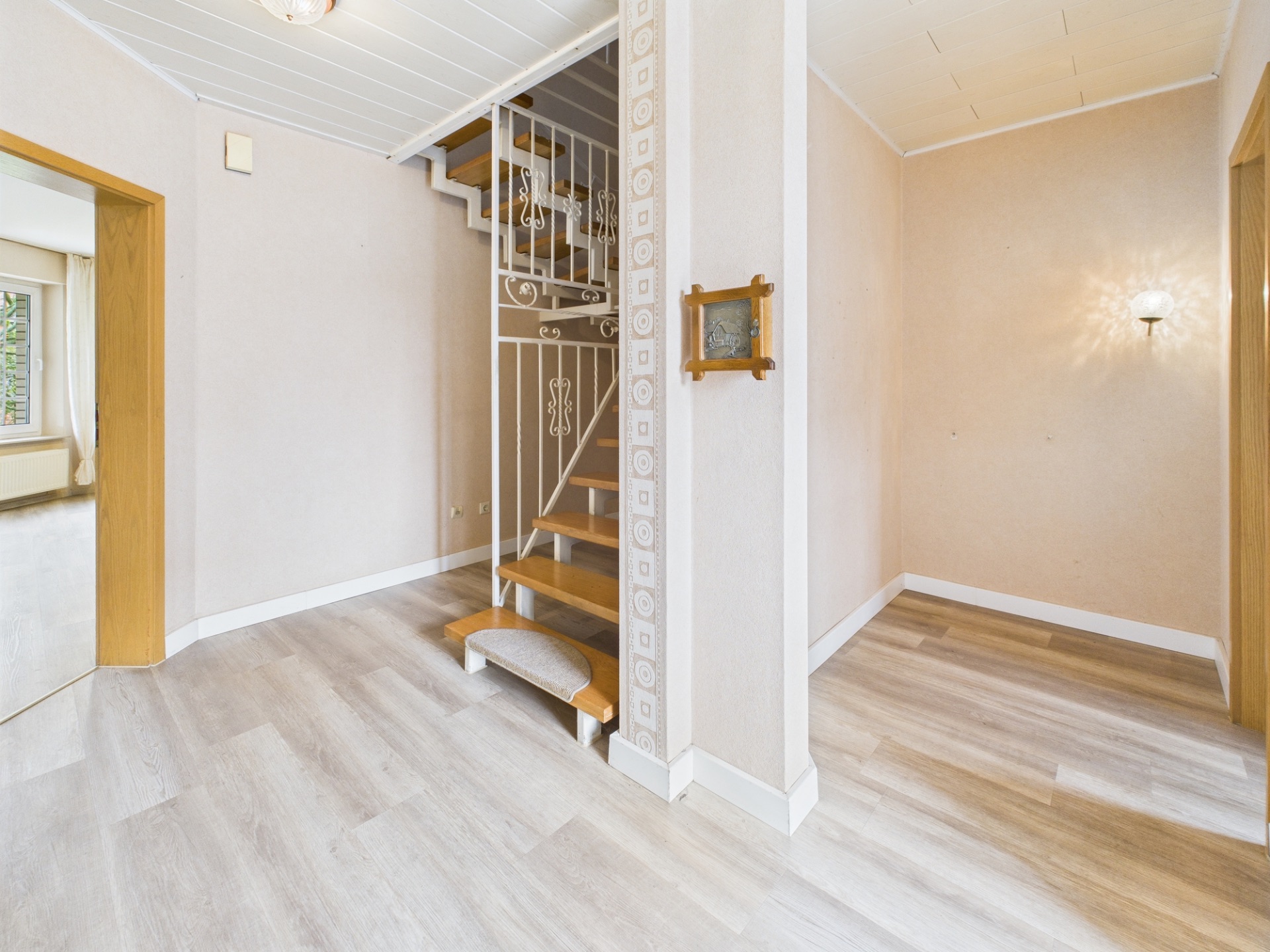 Heller Flur mit Holztreppe und Metallgeländer, hellem Boden und Wandlampe.