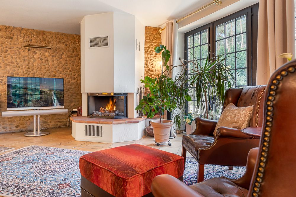 Gemütliches Wohnzimmer mit Kamin, Ledersessel, Teppich, großen Fenstern und Zimmerpflanzen.