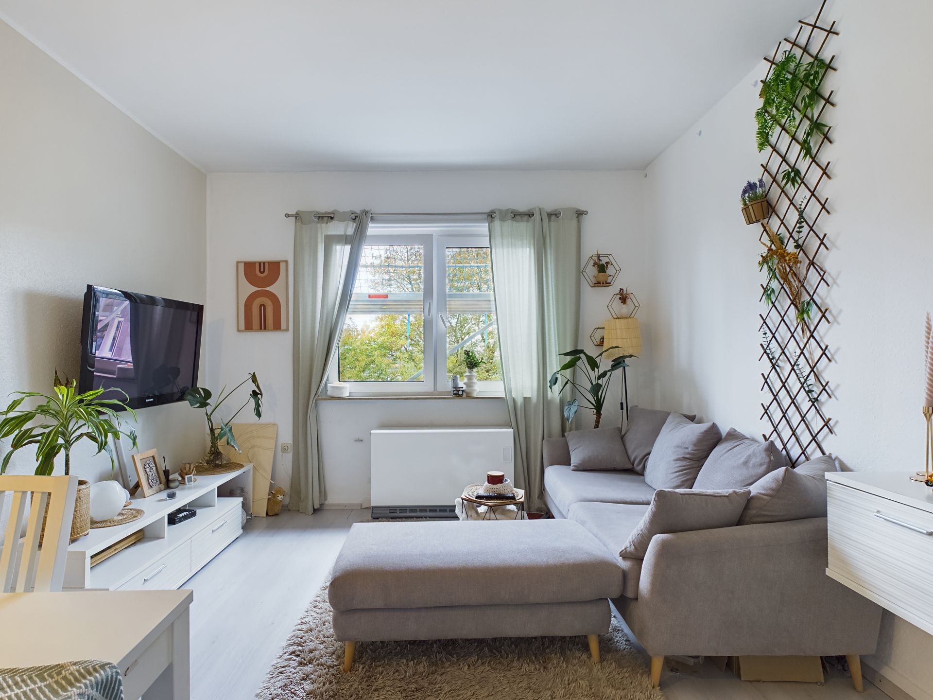 Helles Wohnzimmer mit grauem Sofa, Pflanzen, Wand-TV und Fenster mit Blick ins Grüne.
