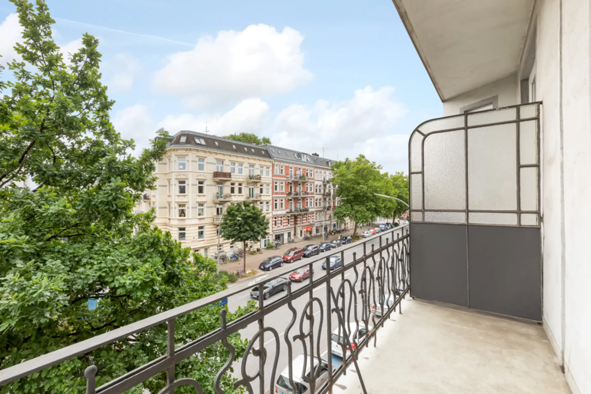 Balkon mit schmiedeeisernem Geländer und Blick auf eine Straße mit Bäumen und Altbauten.