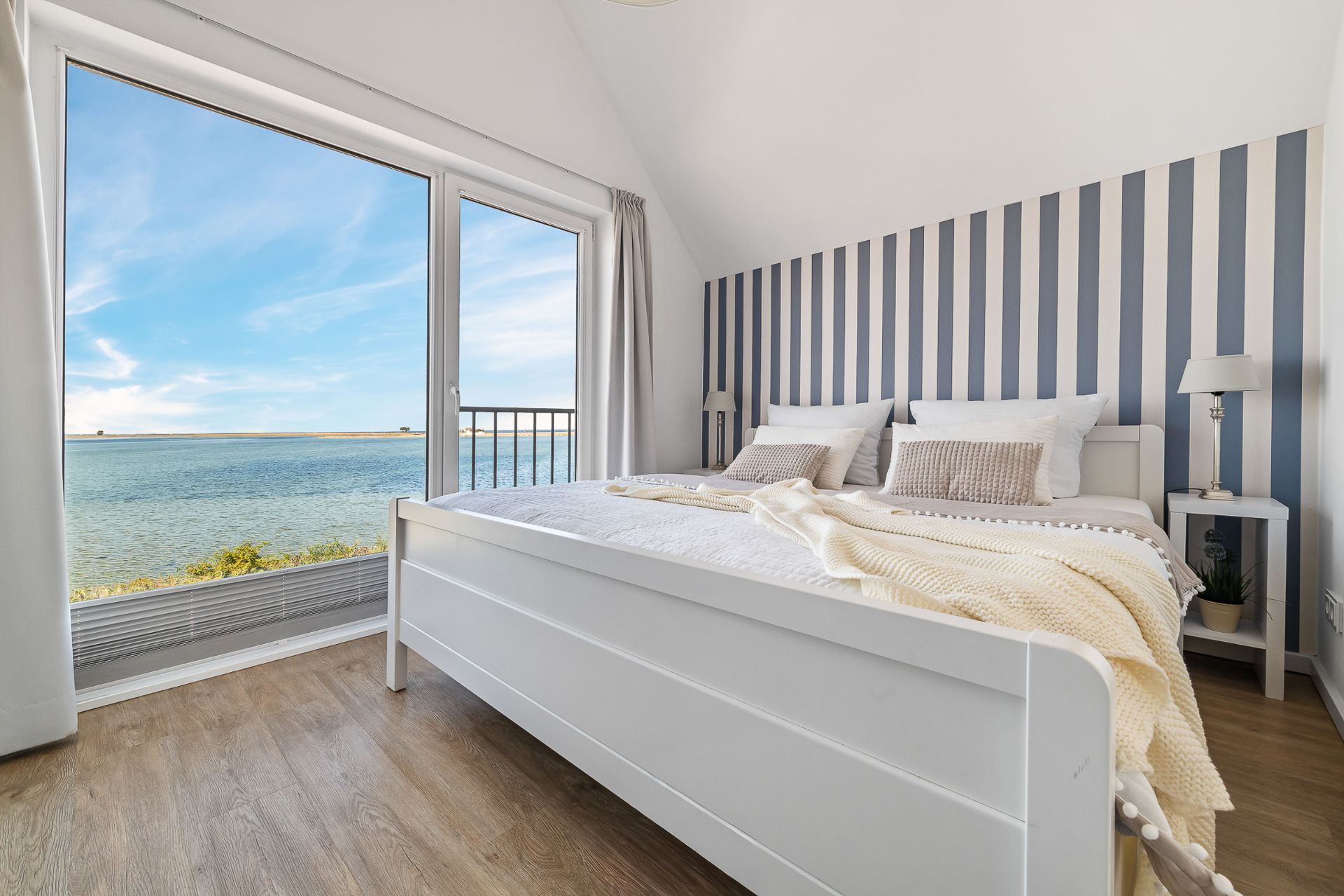 Helles Schlafzimmer mit großem Fenster und Blick auf das Wasser, Doppelbett und gestreifter Akzentwand.