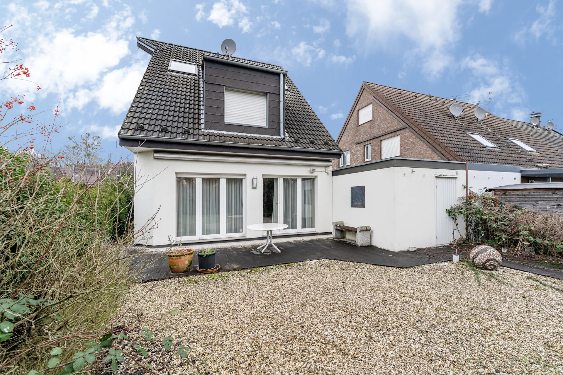 Hinterhof eines Hauses mit Terrasse, Tisch, zwei Blumentöpfen und Kiesfläche.