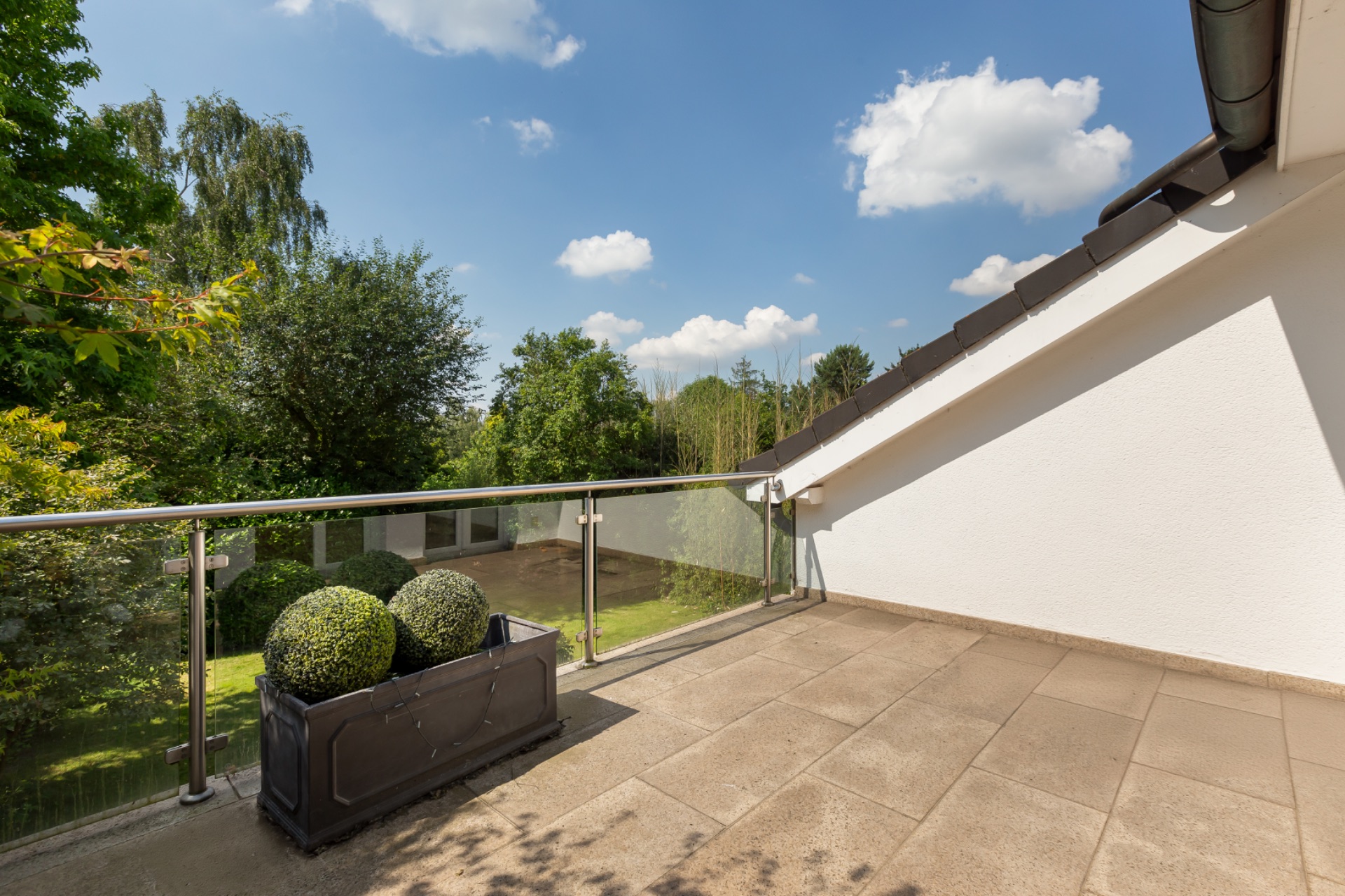 Sonnige Terrasse mit Glasgeländer und Pflanzkübel, Blick auf Garten und Bäume.
