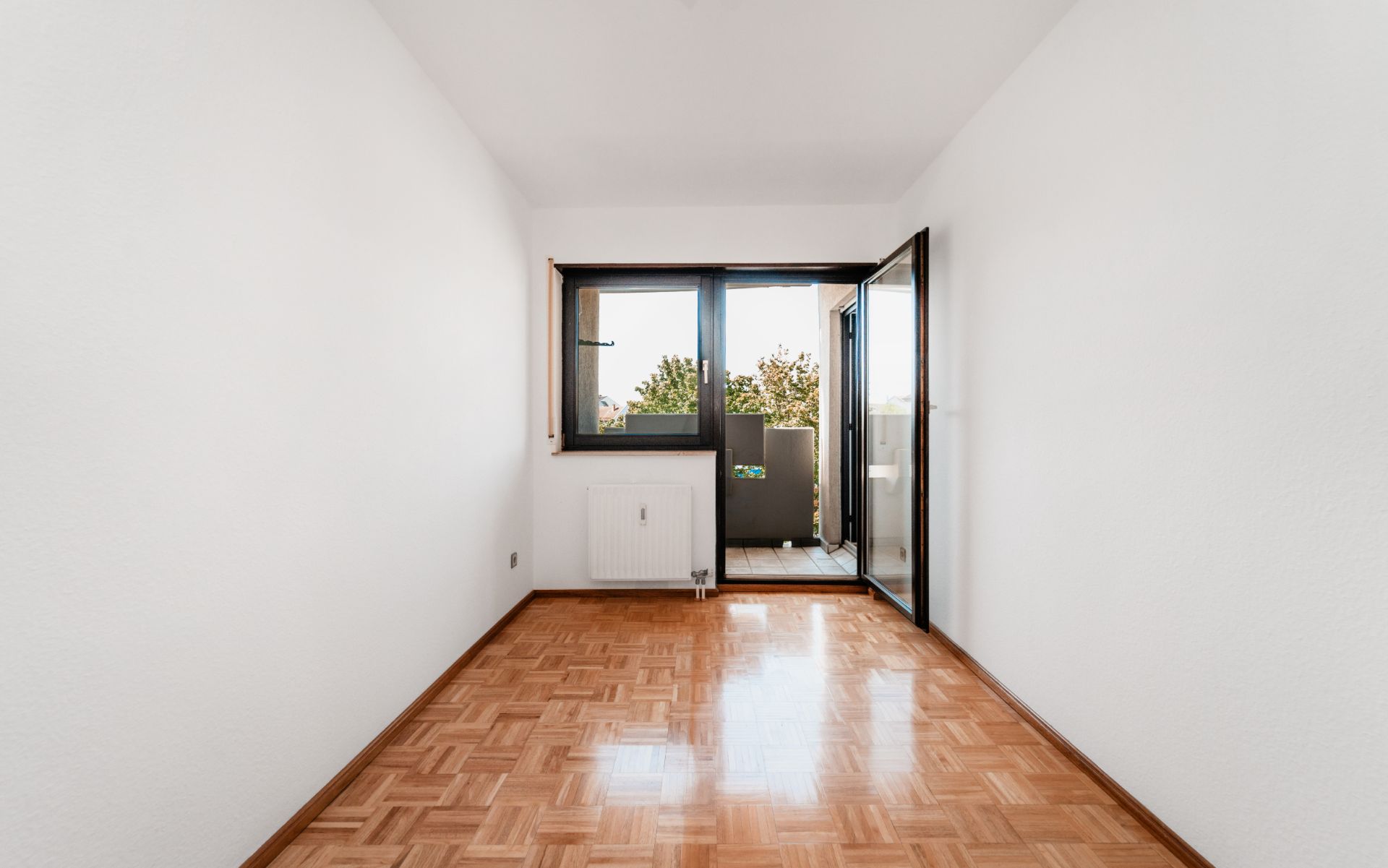 Leerer Raum mit Parkettboden, weiß gestrichenen Wänden und Zugang zu einem Balkon.