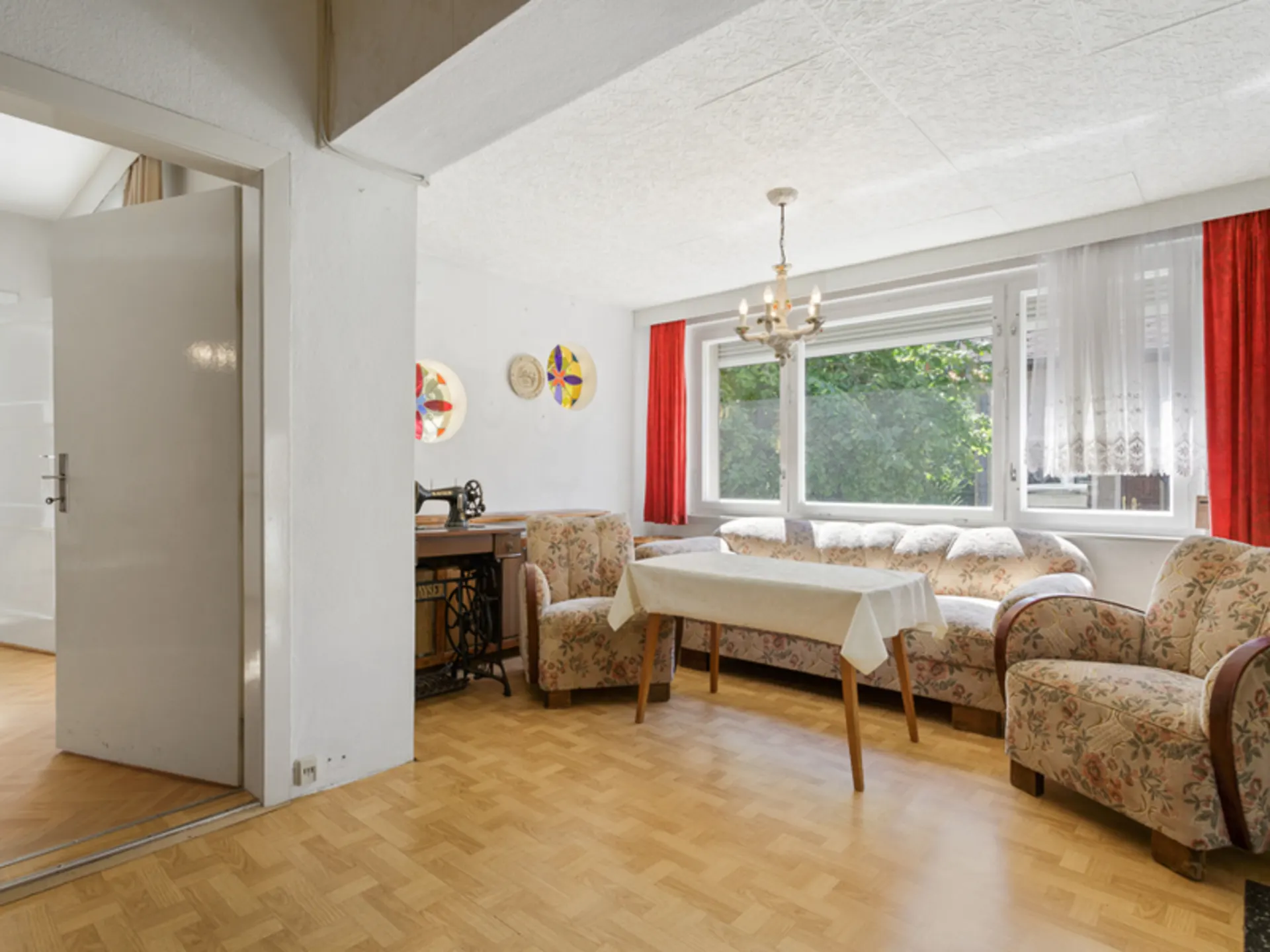 Wohnzimmer mit floralen Polstermöbeln, Esstisch, großer Fensterfront und rotem Vorhang.