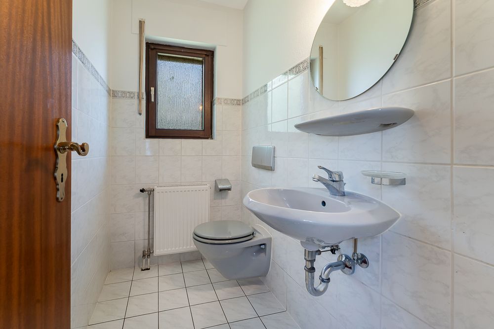 Kleines Badezimmer mit WC, Waschbecken, Spiegel, Fenster und Fliesenboden.