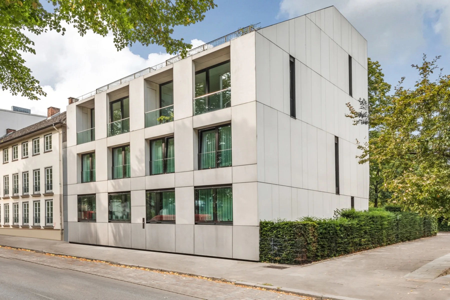 Modernes, eckiges Wohngebäude mit großen Fenstern und hellgrauer Fassade an einer Straße.