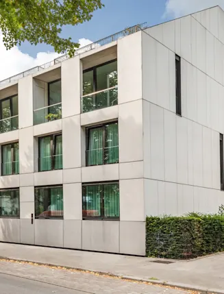 Modernes, eckiges Wohngebäude mit großen Fenstern und hellgrauer Fassade an einer Straße.