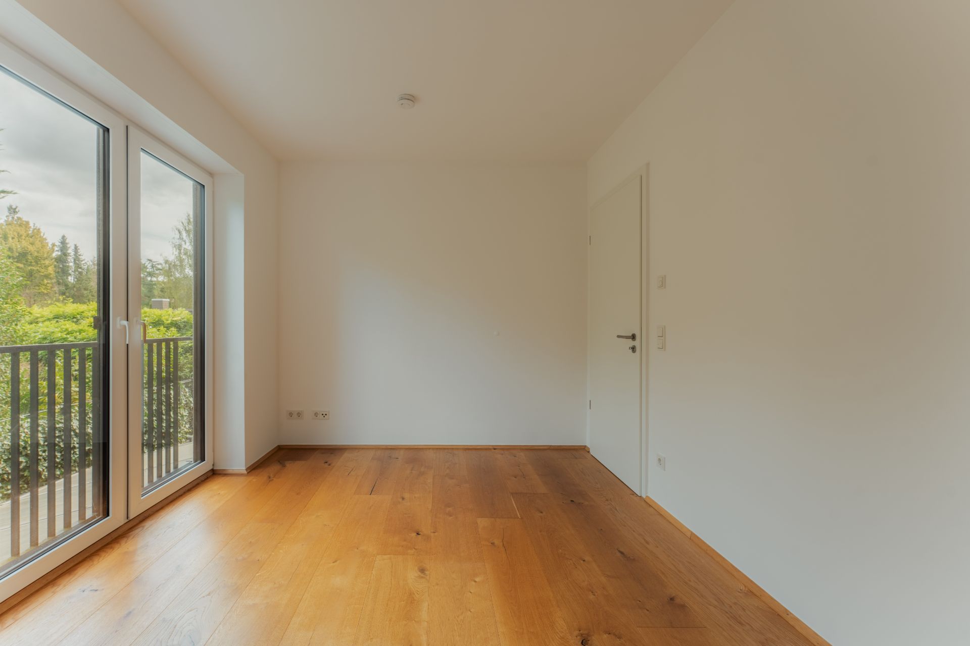 Leerer Raum mit Holzboden, weißen Wänden und großen Fenstern mit Blick auf den Balkon.