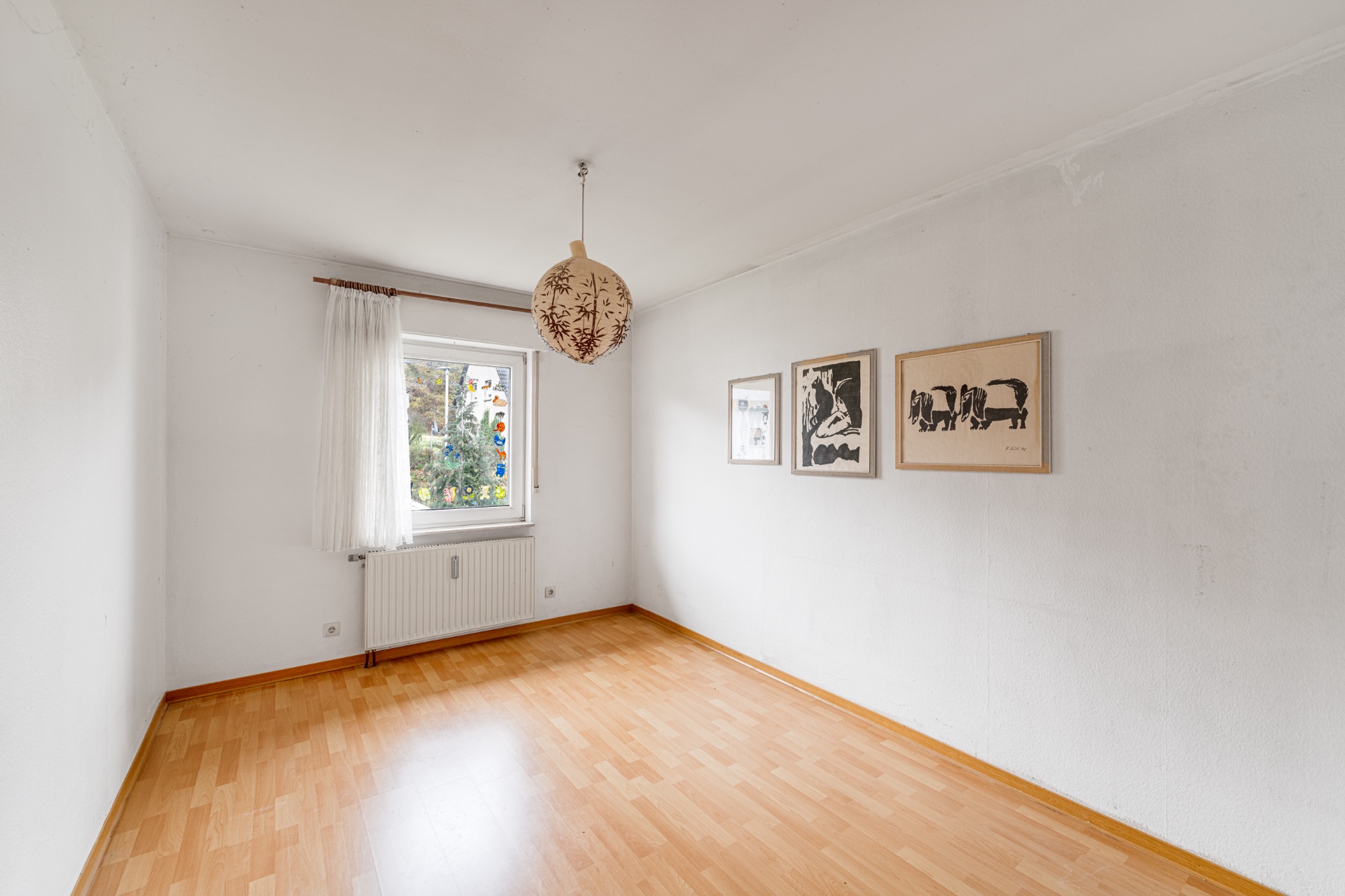 Heller Raum mit Holzfußboden, Fenster, weißem Vorhang und drei Bildern an der Wand.