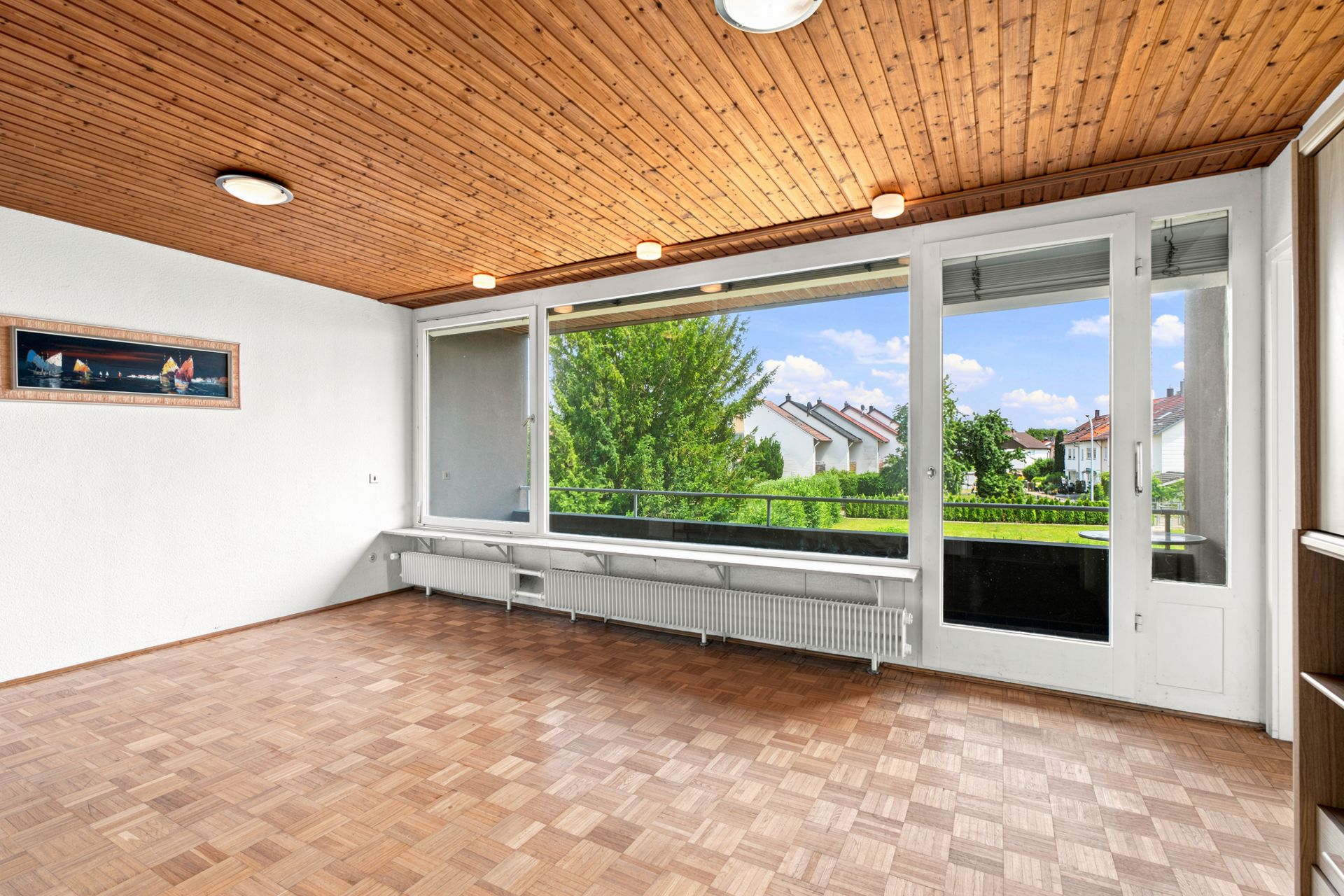 Leeres Wohnzimmer mit Parkettboden, Holzdecke und großen Fenstern mit Blick ins Grüne.