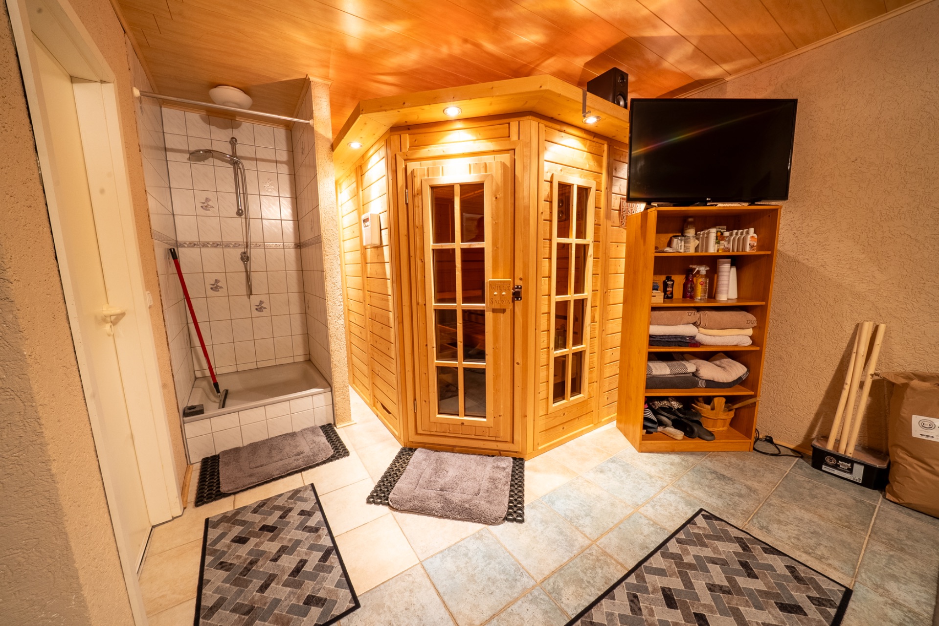 Zimmer mit Holzsauna, Dusche, Regal mit Handtüchern und Fernseher, Fliesenboden, Teppiche.