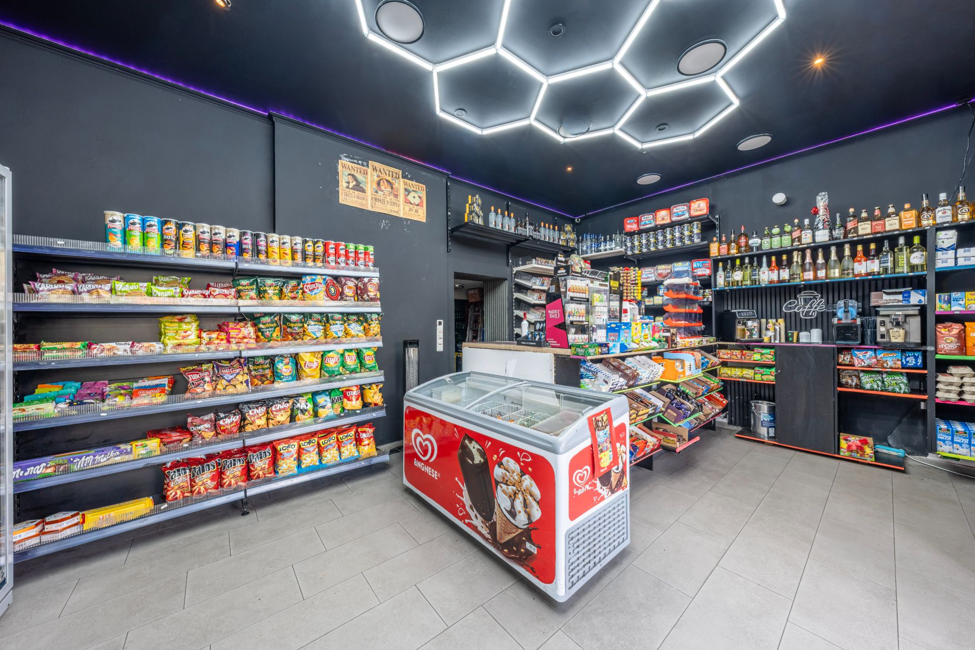 Kleiner Laden mit Regalen voller Snacks, Tiefkühltruhe und beleuchtetem, dunklem Deckendesign.