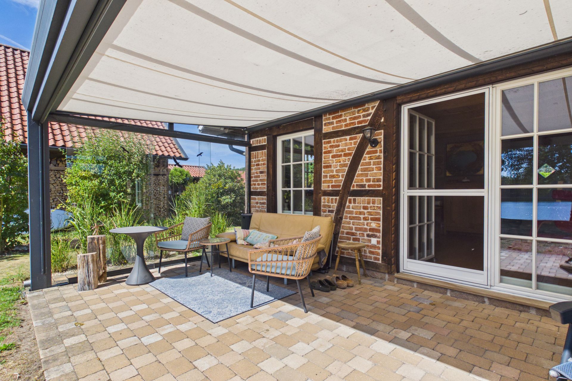 Überdachte Terrasse mit Sitzgruppe vor einem gemauerten Wohnhaus und großem Fenster.