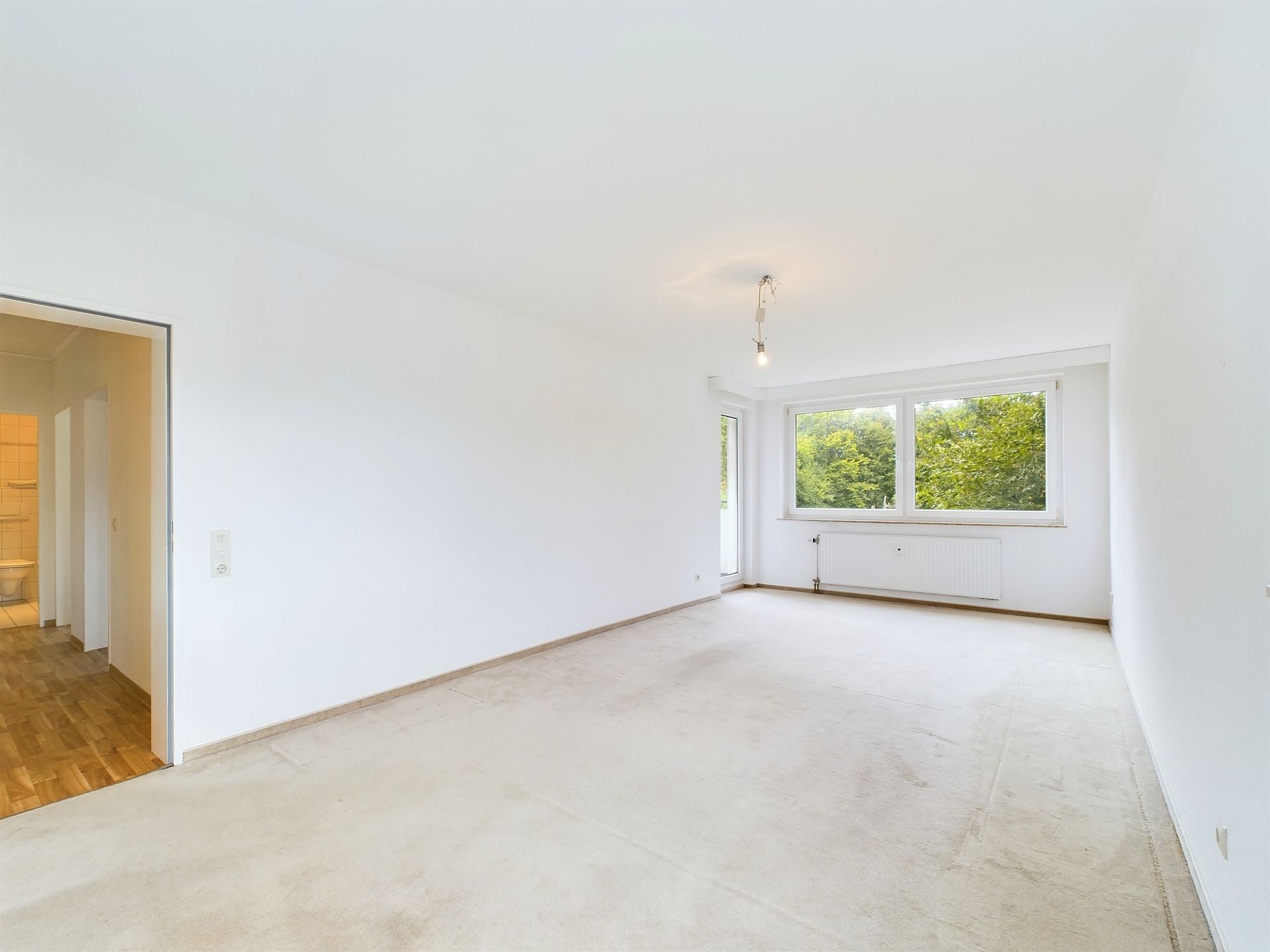 Helles, leeres Zimmer mit großen Fenstern, weiß gestrichenen Wänden und Teppichboden.