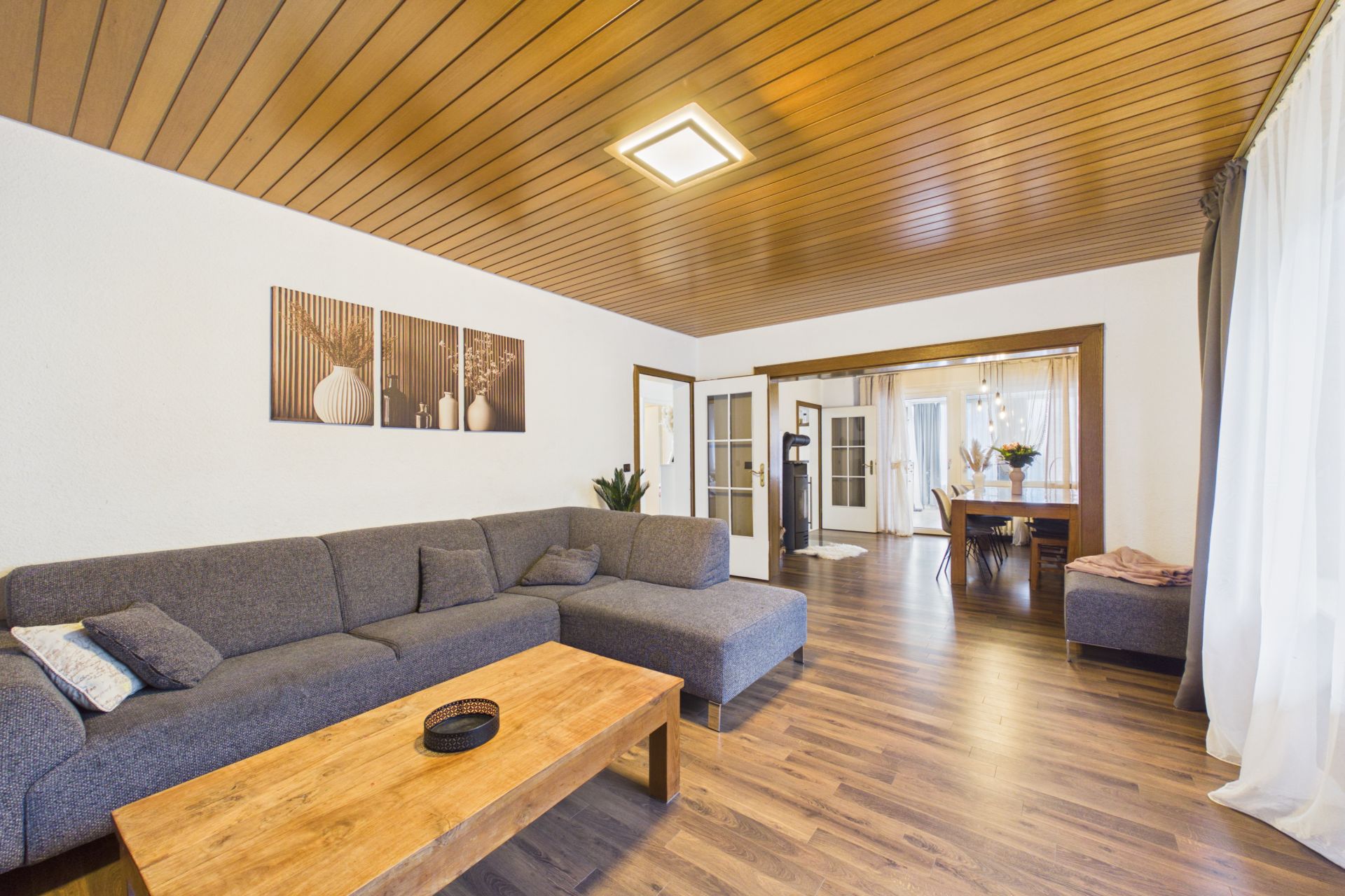 Modernes Wohnzimmer mit grauem Ecksofa, Holztisch und Essbereich im Hintergrund.
