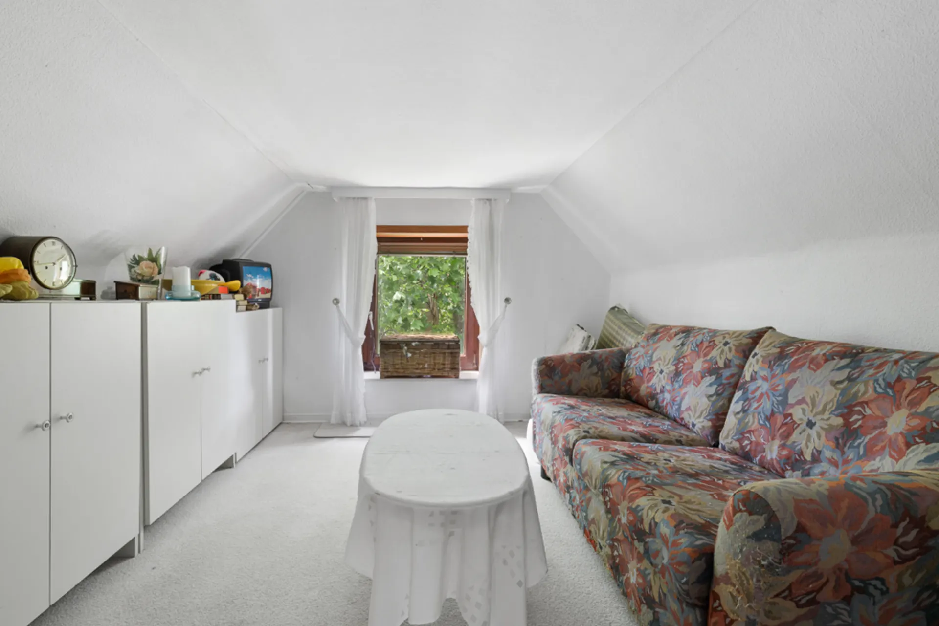 Kleines Wohnzimmer mit bunter Couch, weißem Tisch und Fenster mit Blick ins Grüne.