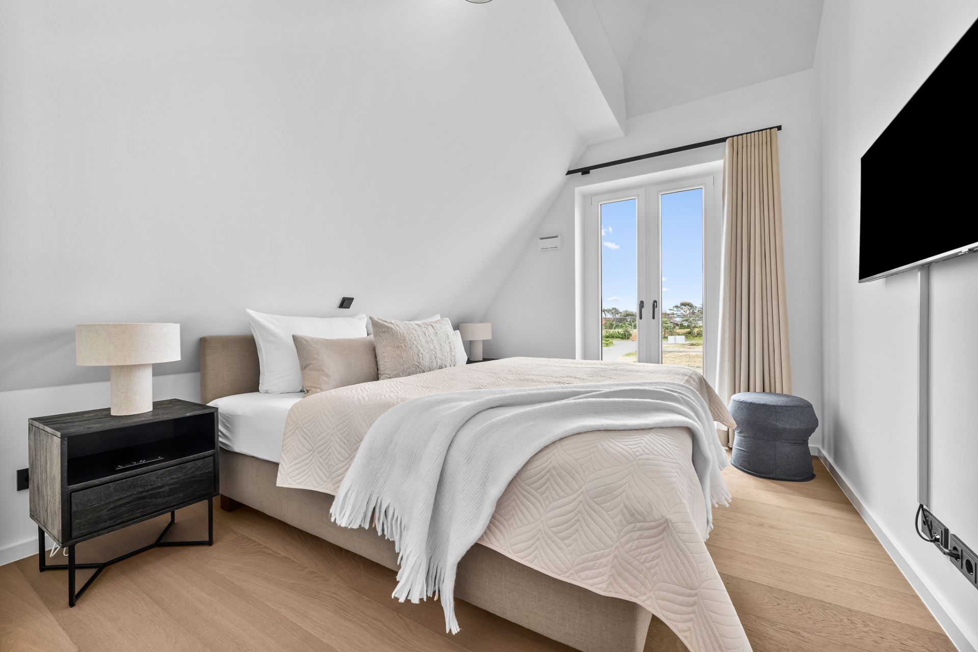 Helles Schlafzimmer mit Doppelbett, Nachttisch, Lampe, TV und Türen zum Balkon.
