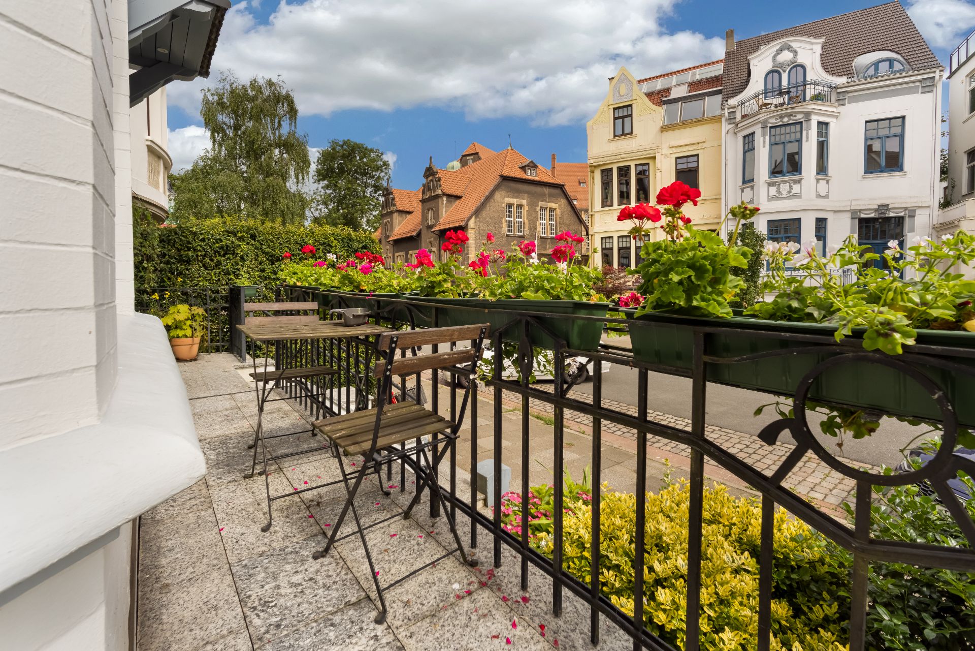 Kleine Terrasse mit Holztisch, Stühlen und Blumenkästen voller roter Blumen, Blick auf Wohnhäuser.