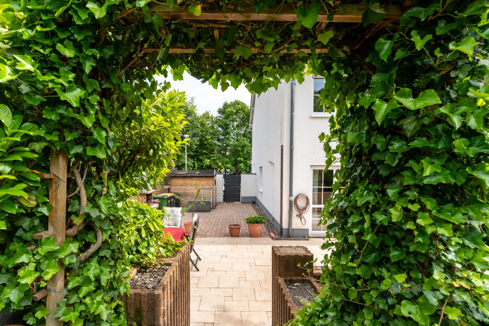 Durch einen mit Efeu bewachsenen Torbogen sieht man auf eine Terrasse und ein weißes Wohnhaus.