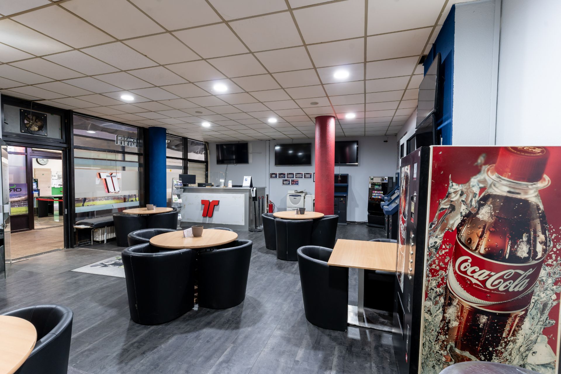 Heller Raum mit mehreren runden Tischen, schwarzen Sesseln und einem großen Coca-Cola-Automaten.