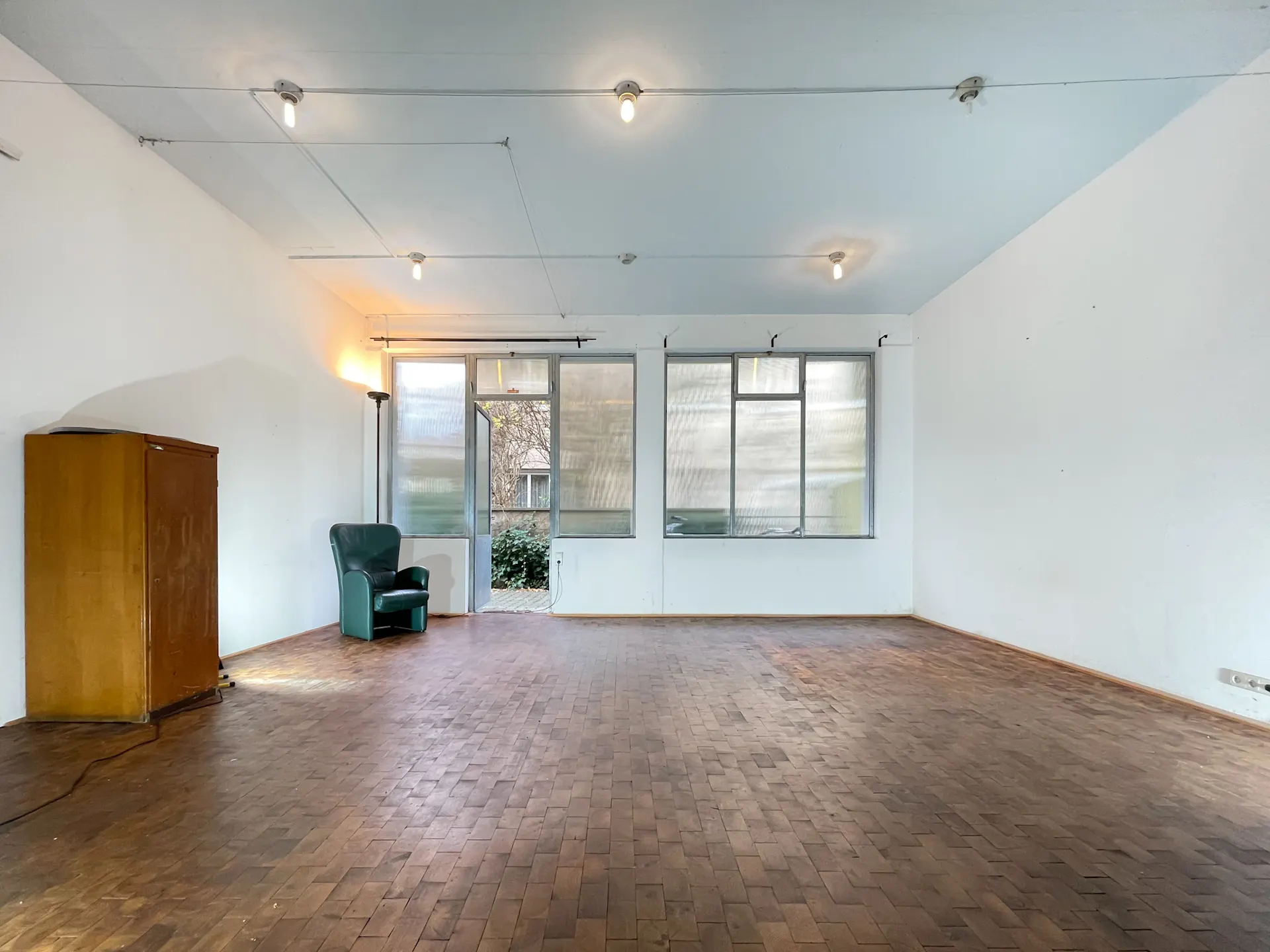 Leerer Raum mit Holzfußboden, grünem Sessel, Schrank und großen Fenstern.