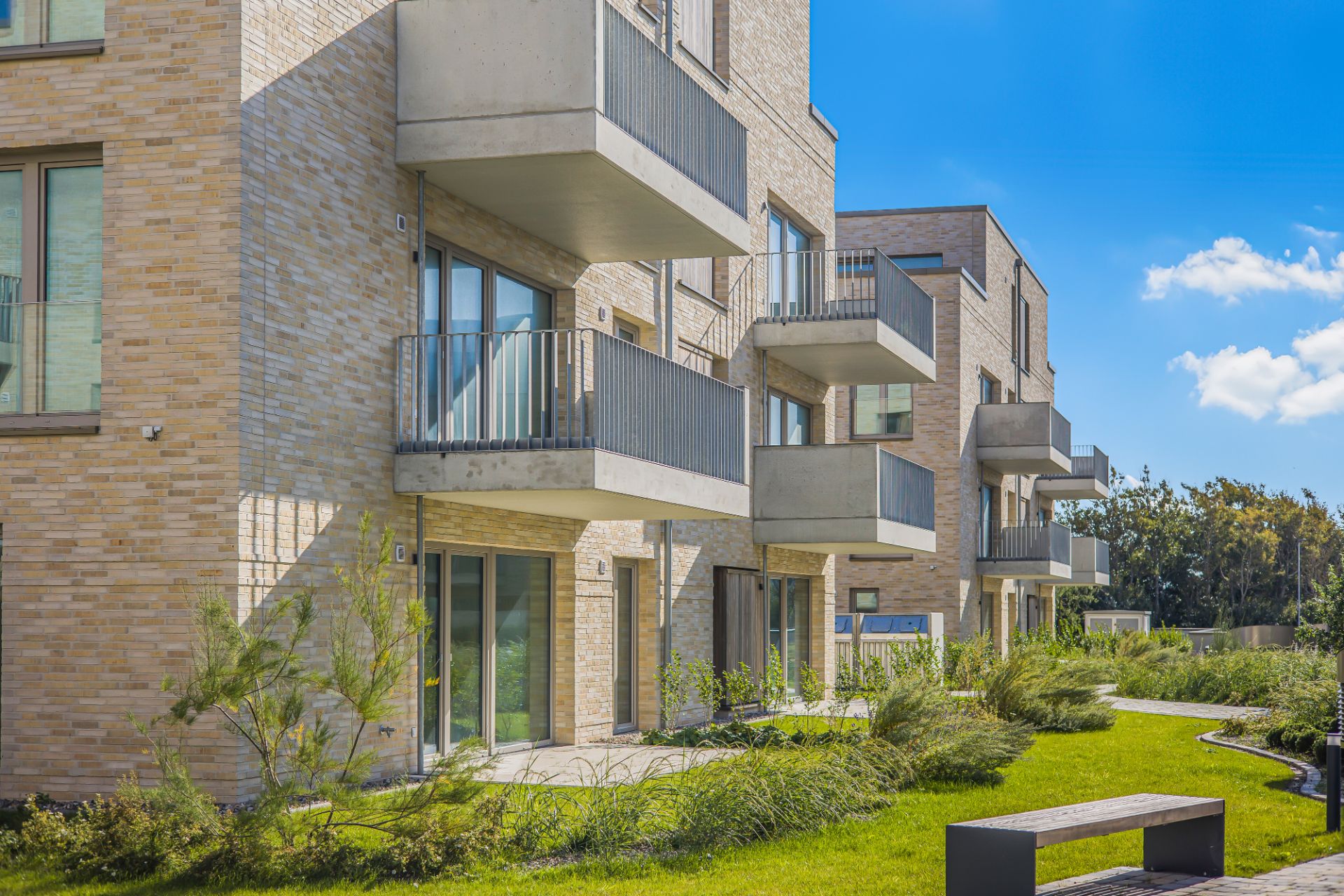 Moderne Wohngebäude mit Balkonen und großen Fenstern, davor gepflegter grüner Garten mit Bank.