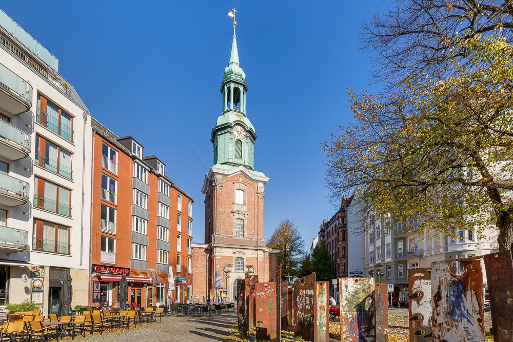 Kirchturm mit grünem Dach zwischen Wohnhäusern, vorne Bänke und Bäume.