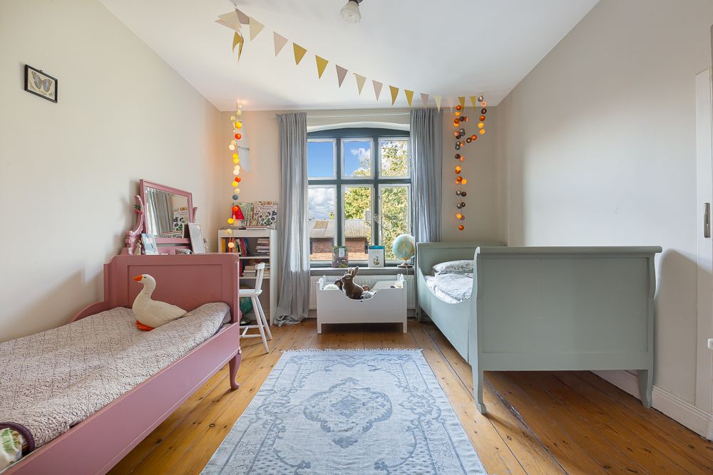 Helles Kinderzimmer mit zwei Betten, Schreibtisch, Fenster und bunten Girlanden.