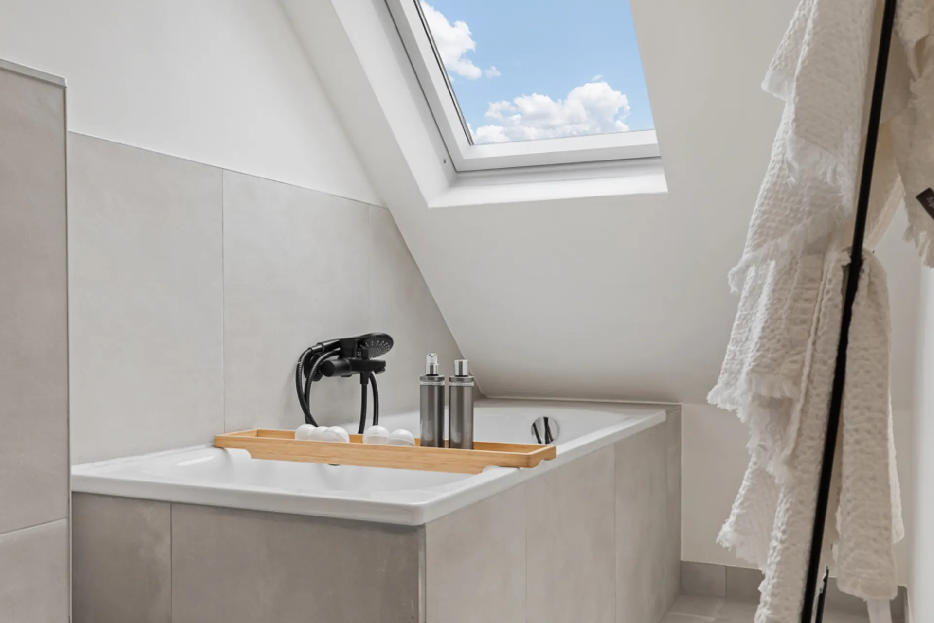 Modernes Bad mit Badewanne unter einem Dachfenster, Ablagebrett und hellen Fliesen.