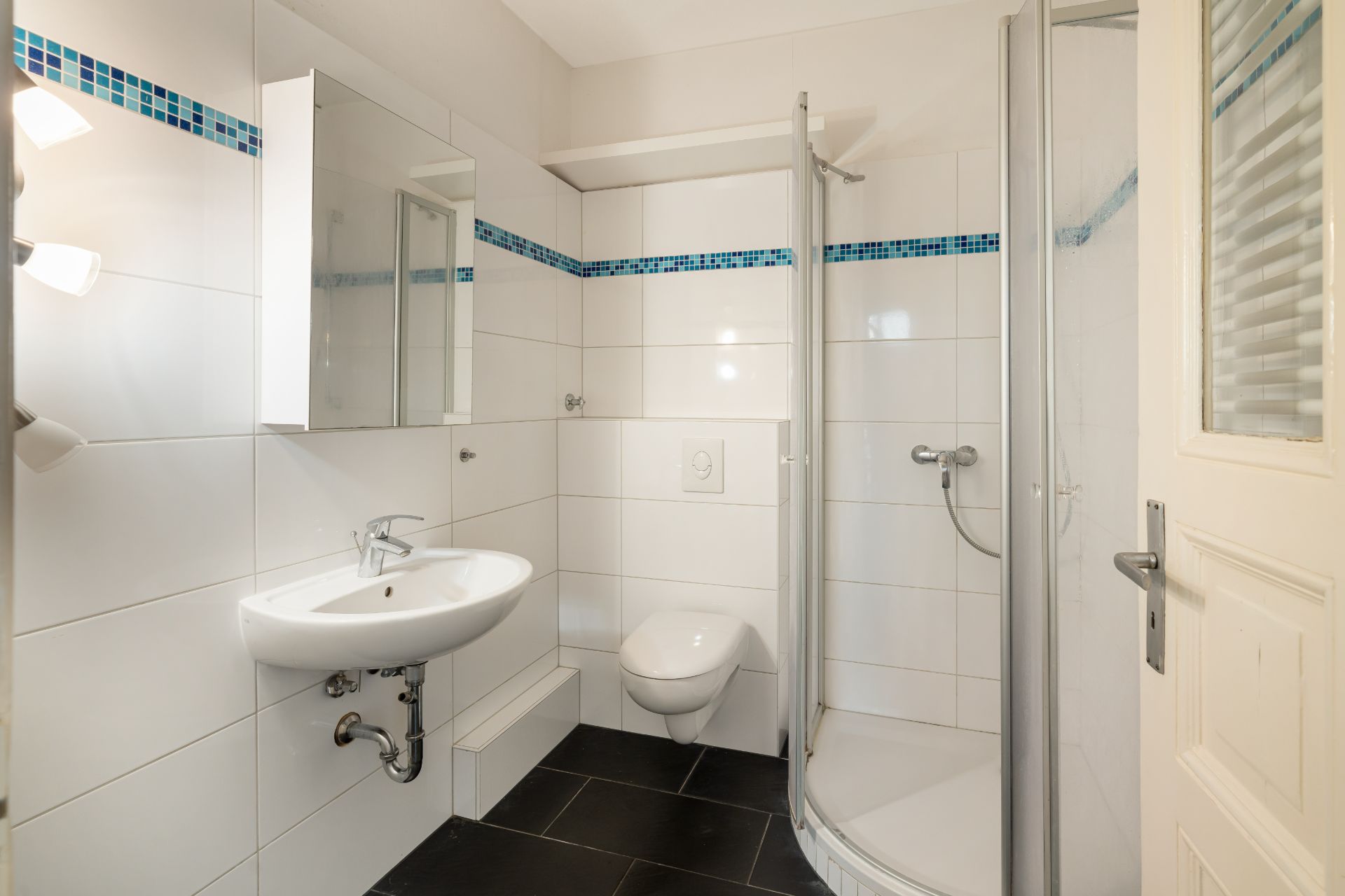 Modernes Badezimmer mit Dusche, Waschbecken, Spiegel und Toilette, weiße Fliesen mit blauem Akzentband.