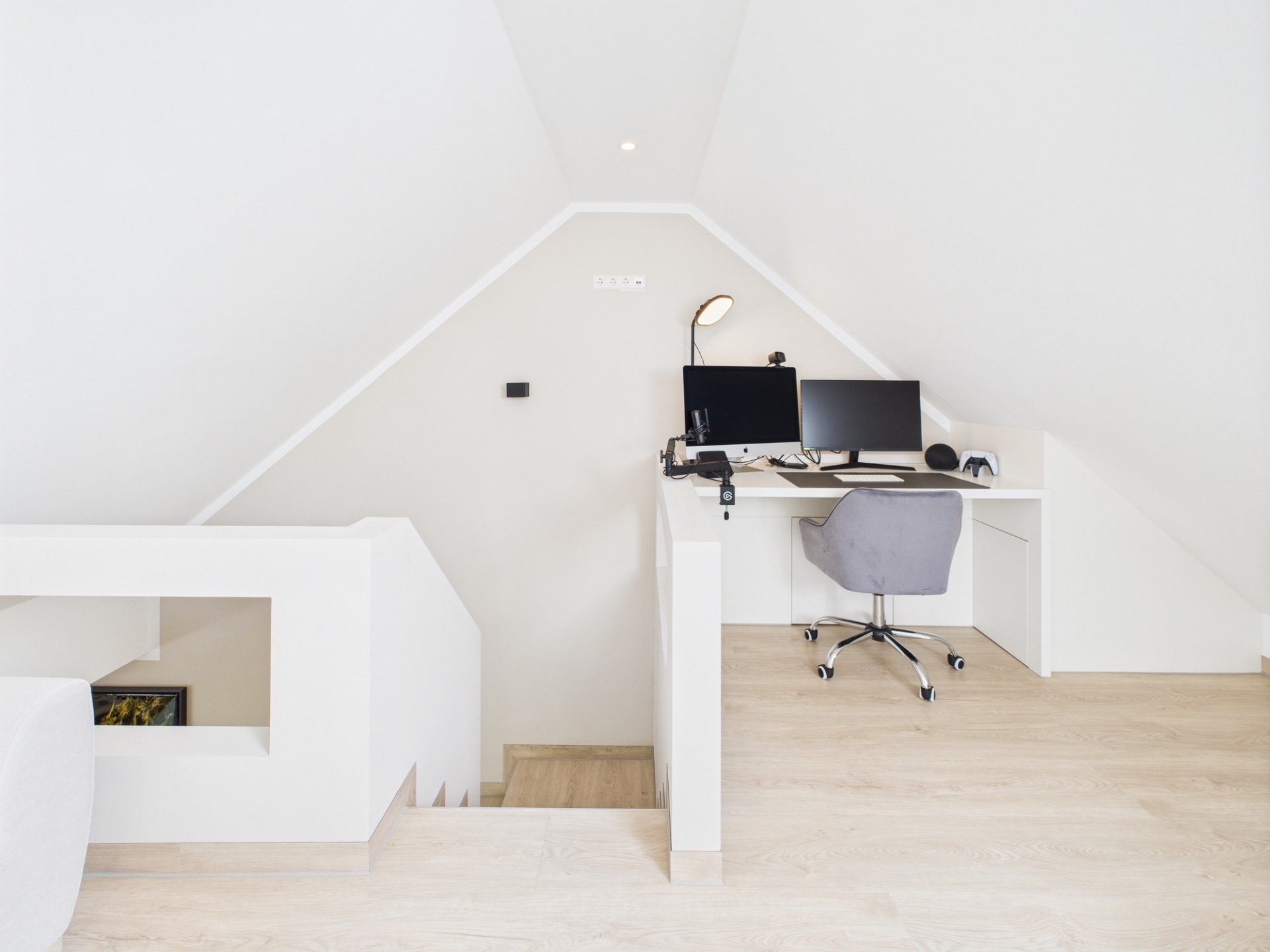 Helles Dachzimmer mit Schreibtisch, zwei Monitoren und einem Bürostuhl.