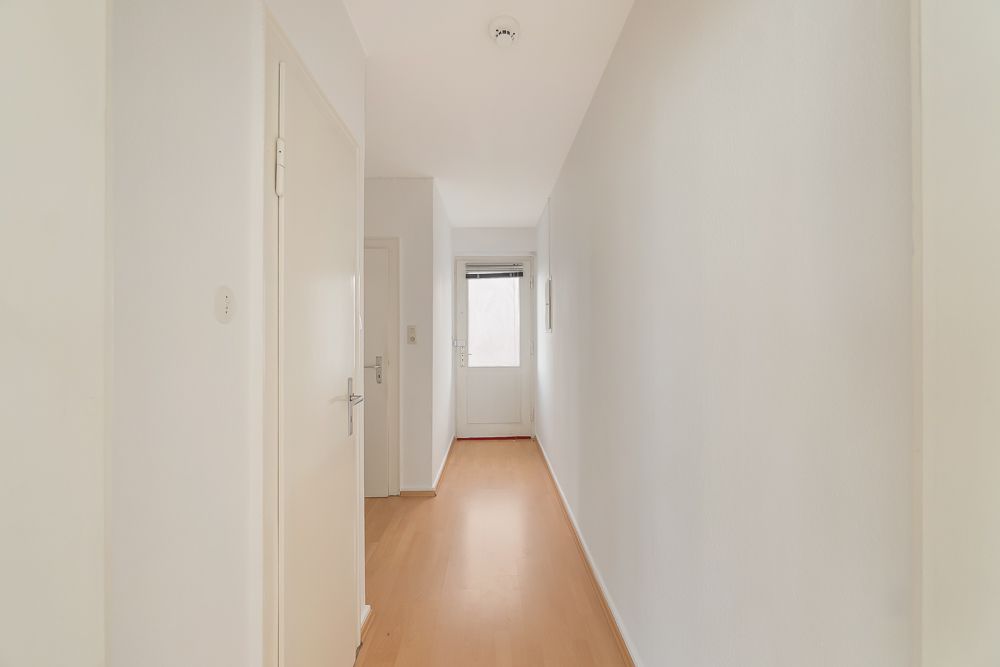 Heller Flur mit Holzboden, weißen Wänden und Türen, am Ende eine Tür mit Fenster.