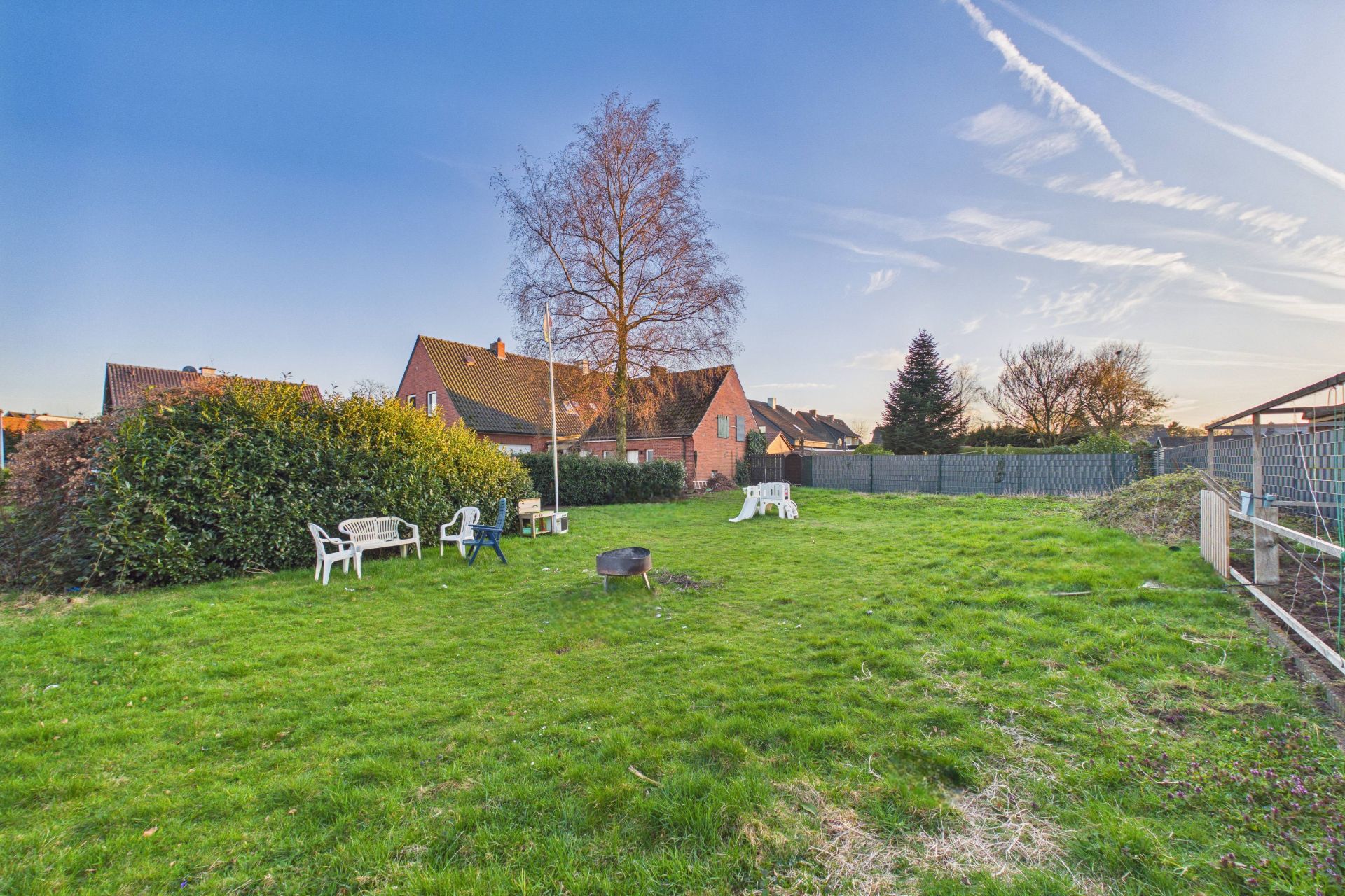 Grüner Garten mit Rasen, Gartenstühlen, Feuerstelle und Blick auf Nachbarhäuser.